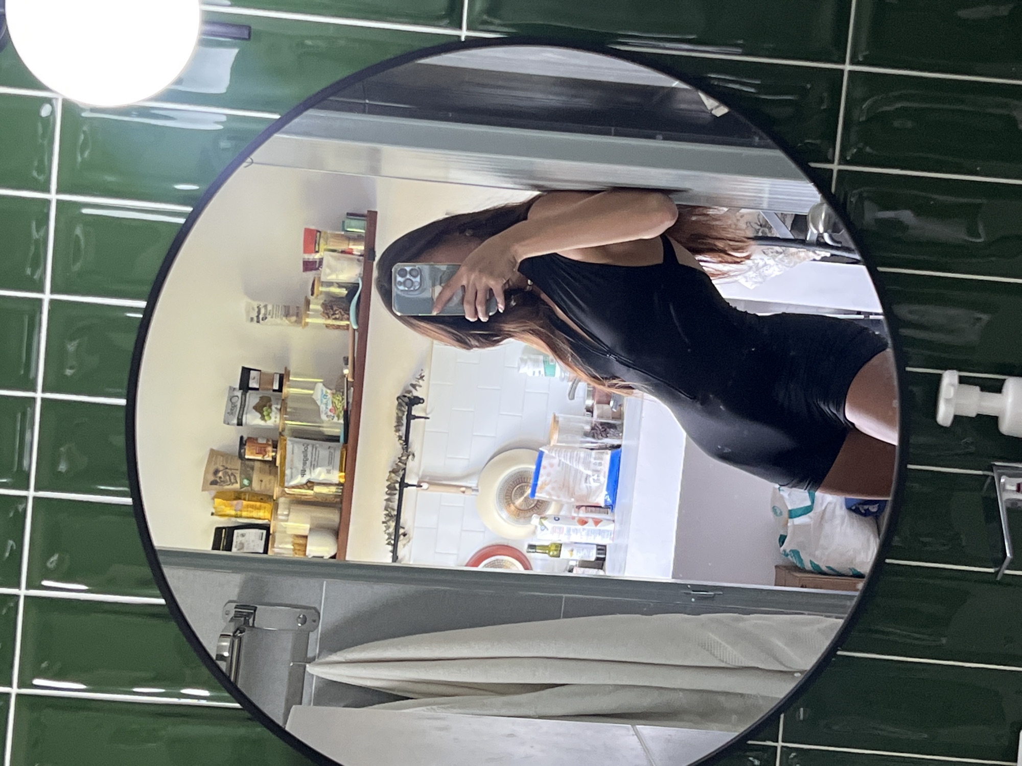 A mirror reflects a woman taking a selfie in a bathroom. The bathroom features green tiled walls and a light fixture on the ceiling. The woman is wearing a black outfit. Behind her in the reflection is a kitchen area with shelves stocked with various items, cooking utensils, and food products. A towel is also visible in the reflection. The location is Unknown, Unknown.