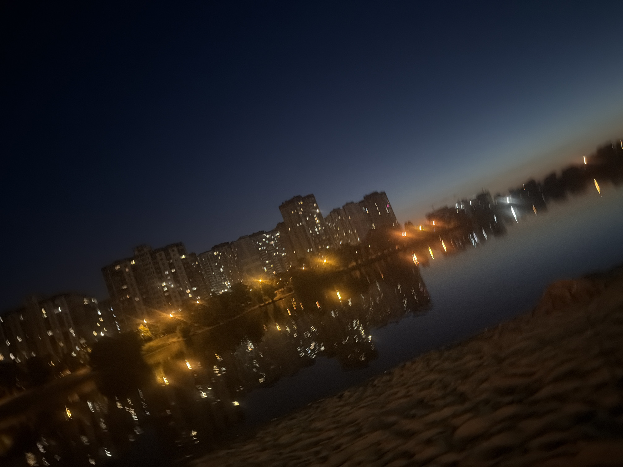 This is a tilted, blurred, nighttime photograph of a city skyline reflected in water. The scene appears to be in Kyiv, Ukraine, given the characteristic Soviet-era apartment blocks that dominate the background. Tall, multi-story residential buildings with numerous lit windows are visible. The buildings are clustered together and stretch across the mid-ground of the image. The dark sky above transitions to a lighter hue near the horizon, suggesting either early morning or late evening twilight. The water in the foreground reflects the lights of the buildings and streetlights, creating elongated streaks of light that are distorted by motion or camera shake. The bank of the water body is visible in the lower right corner, with a textured surface that appears to be made of stone or paving. There are no people or specific identifiable objects or actions depicted. The overall impression is one of an urban environment at night, possibly a residential district situated near a body of water like a lake or river.