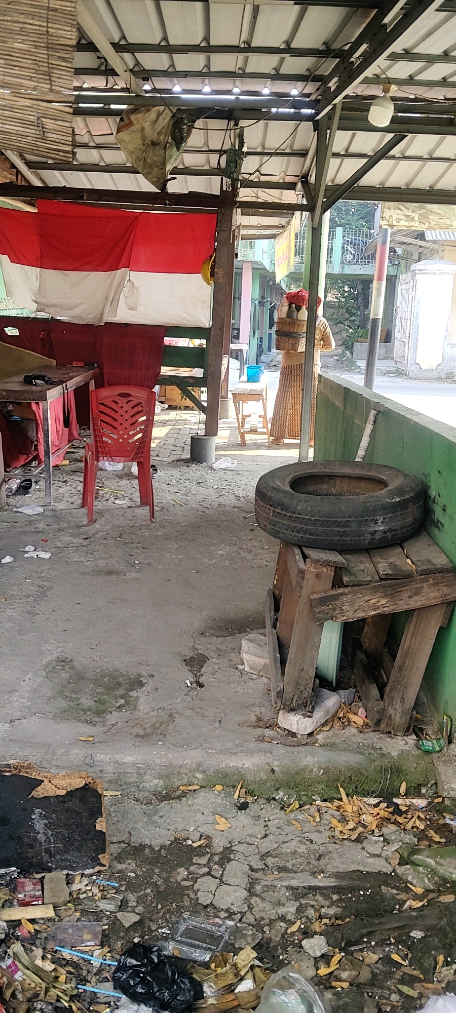The image depicts an outdoor scene in Mekarmukti, Indonesia, characterized by a covered area with a corrugated metal roof supported by metal beams. The area appears to be a makeshift stall or shop. A large Indonesian flag, red and white, is prominently displayed, hanging from the structure.
In the foreground, the ground is a concrete surface, littered with debris including plastic bags, food packaging, leaves, and what appears to be a broken piece of wood with the Indonesian flag draped over it. A red plastic chair is positioned in the center of the space. To the right, a worn wooden stand holds an old tire.
Further back, a woman is visible carrying a basket on her head, containing items that appear to be red flowers or food. She is walking away from the camera towards a street scene, where other buildings and a motorcycle are visible. The overall impression is one of a functional but somewhat unkempt outdoor commercial space in a local Indonesian setting.
