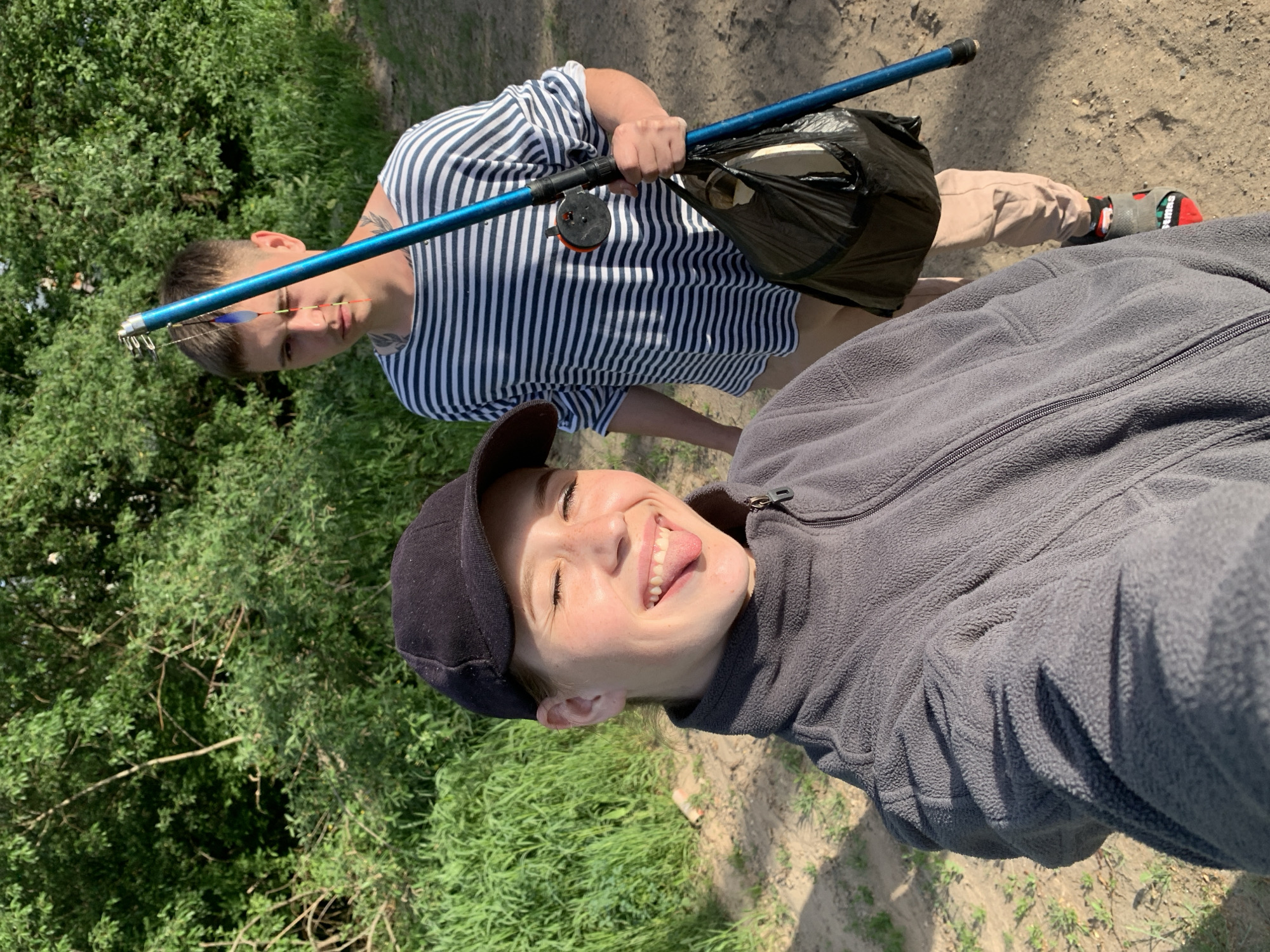 Two people pose outdoors, one holding a blue fishing rod. The foreground person wears a black cap and a gray fleece jacket, sticking out their tongue and smiling. The background person, positioned behind and to the left, wears a striped shirt, tan pants, and red and black shoes, holding the fishing rod. A black bag is attached to the rod. The ground is dirt and grass, with green trees and bushes in the background. The location is likely Фоки, Russia.