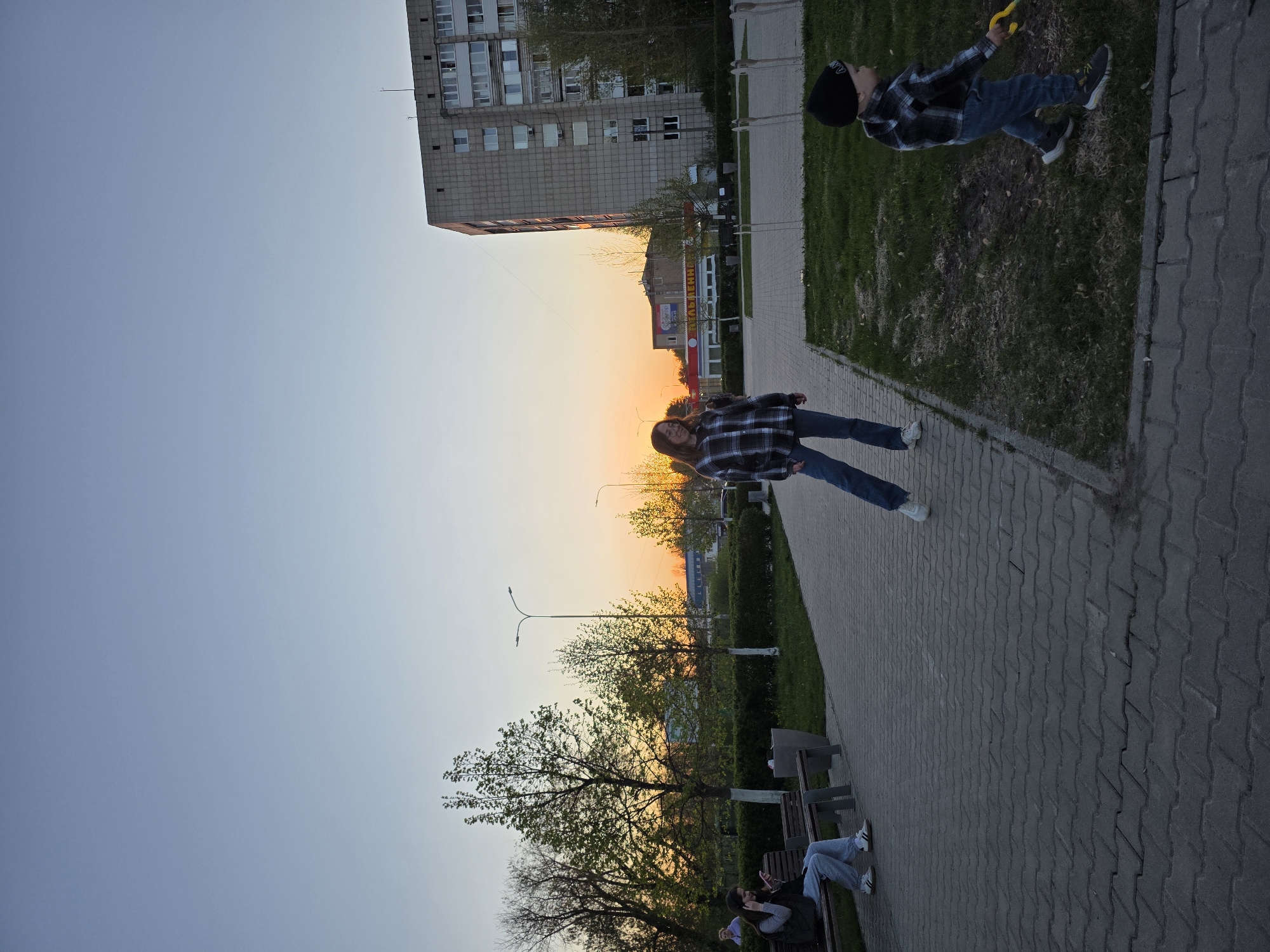 **Scene:** A pedestrian walkway in Chaikovsky, Russia, at sunset.
**People:**
* A young woman wearing a blue and black plaid shirt and blue jeans walks along the pavement.
* A young boy, wearing a black beanie, a blue and black plaid shirt, and blue jeans, holds a yellow object and plays in a grassy area adjacent to the path.
* Two individuals sit on a park bench.
**Environment:**
* A paved walkway with park benches along the side.
* Grass patches bordering the walkway.
* Trees in the background with a sunset sky.
* Tall apartment buildings and other buildings in the distance.
* Street lamps are present.
**Objects:**
* Park benches
* Street lamps
* Buildings
* Yellow object held by the child.