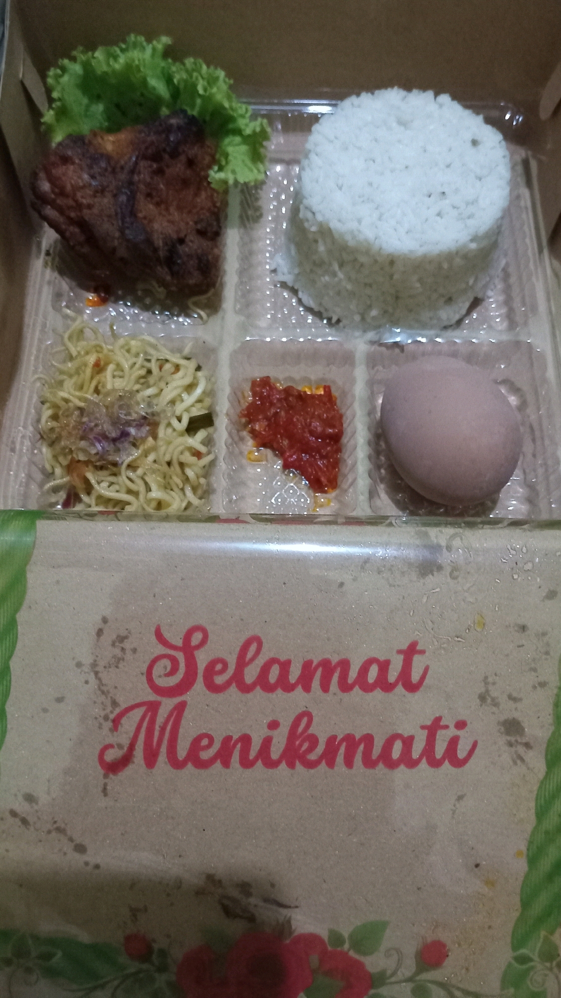 The image shows a bento box meal containing fried chicken, rice, noodles, chili sauce, and a hard-boiled egg. The bento box is placed on a surface that appears to be cardboard with text that reads "Selamat Menikmati", which translates to "Enjoy your meal" in Indonesian. The background suggests an indoor setting, possibly a restaurant or food stall. The lighting is dim and appears to be artificial. There are no people or animals visible in the image. The meal is presented in individual compartments within the bento box, suggesting it is a pre-packaged meal. The bento box itself is made of clear plastic with a cardboard lid. The overall impression is a simple, ready-to-eat meal. Based on the text and the common presentation of such meals, it is plausible that this bento box was purchased in Randuagung, Indonesia.