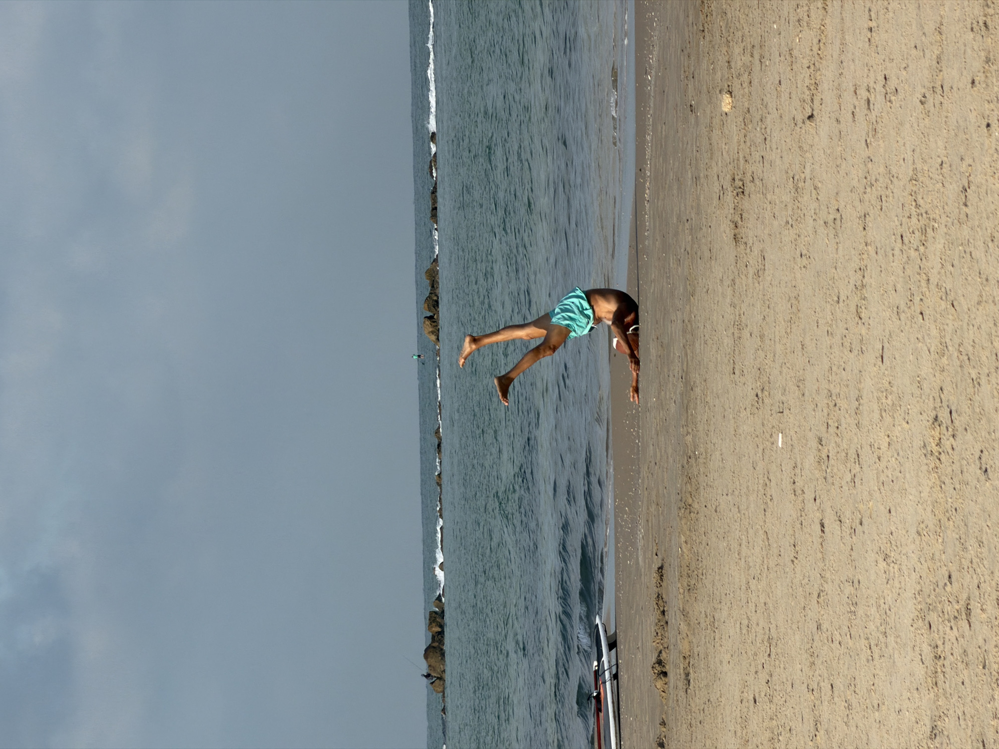 A person in turquoise shorts is diving headfirst into the ocean at a beach. The scene is in Bat Yam, Israel, with a calm, gray sky overhead, a sandy beach on the right, and the Mediterranean Sea to the left. A surfboard is partially visible on the sand near the water's edge. A rock barrier is positioned along the sea with small waves breaking.