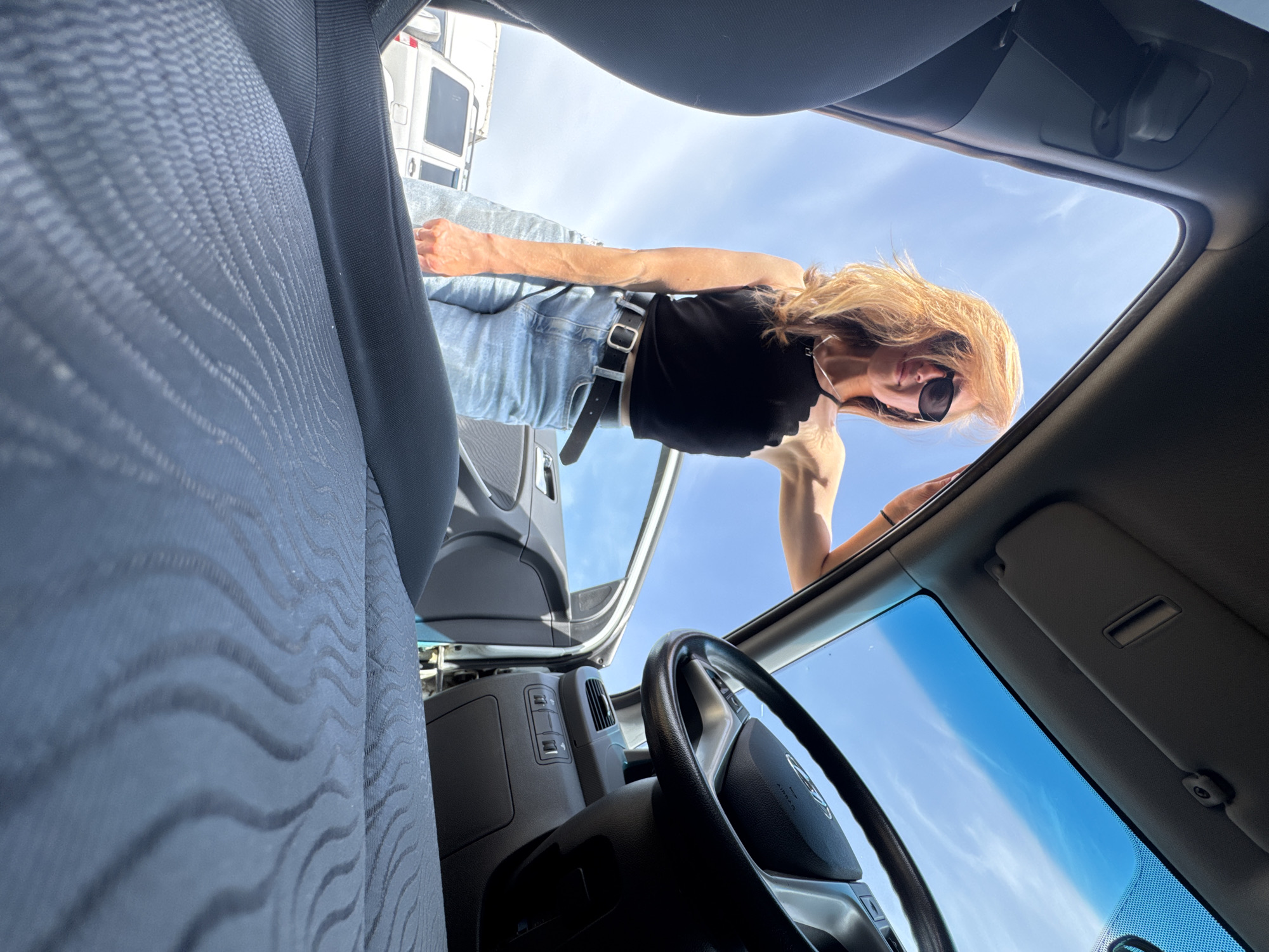 A woman with blonde hair and sunglasses, wearing a black top and jeans with a black belt, is looking down into the open sunroof of a car. The interior of the car, including a seat, steering wheel, and dashboard, is visible. Through the sunroof, the sky and a white vehicle in the background can be seen. The location is Chaikovsky, Russia.