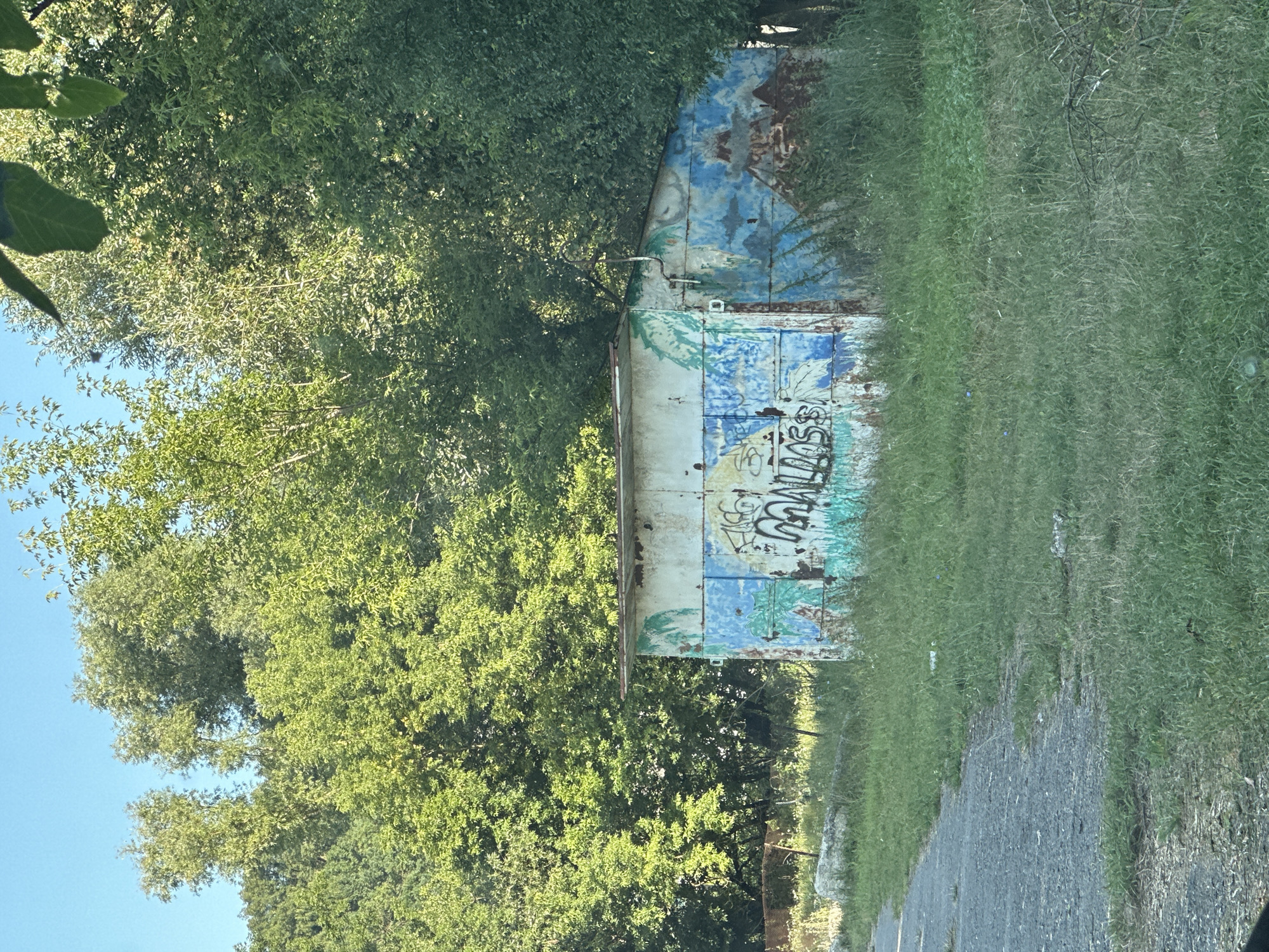 A small, rusted, metal shed with graffiti is situated in a grassy, overgrown area next to a paved path. The shed has a corrugated metal exterior, painted with blue and white sections and what appear to be faded mural designs, possibly depicting palm trees and mountains. Black graffiti tags are visible on the front of the shed. The surrounding environment consists of tall green grass, some weeds, and a dense background of lush green trees under a clear blue sky. The scene appears to be in an outdoor, rural or semi-rural setting, possibly on the outskirts of Makiv, Ukraine.
