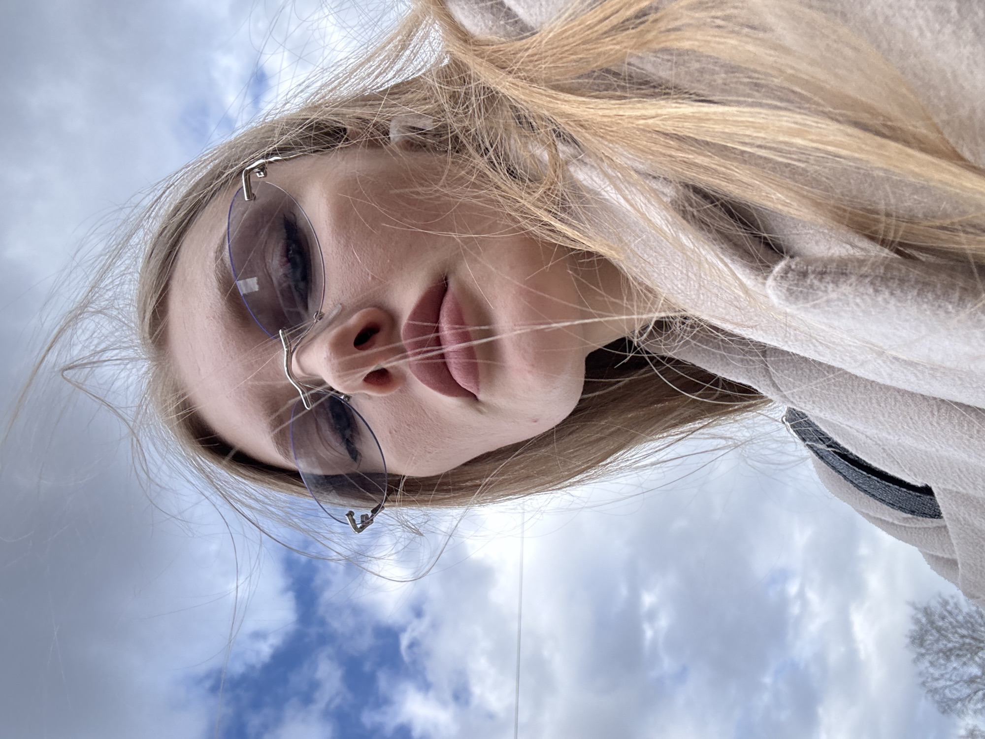 A close-up, low-angle shot captures a young person, likely female, with long, flowing light blonde hair. The subject is wearing modern rimless sunglasses with subtle silver metallic frames, through which their eyes and eyebrows are partially visible. Their lips are slightly parted, creating a soft, casual expression. They are dressed in a light-colored garment, possibly a beige or off-white fleece jacket or hoodie, with a hint of dark lining or trim visible on the shoulder.
The perspective is upward, with the cloudy sky forming the primary background. Patches of blue sky are visible between areas of white and grey clouds, indicating a partly cloudy or clearing overcast day. In the lower right corner, the silhouette of bare tree branches against the sky suggests a colder season, such as late autumn, winter, or early spring. The image appears to be a casual outdoor portrait or selfie, taken during daytime in Perm, Russia. No visible text is present.