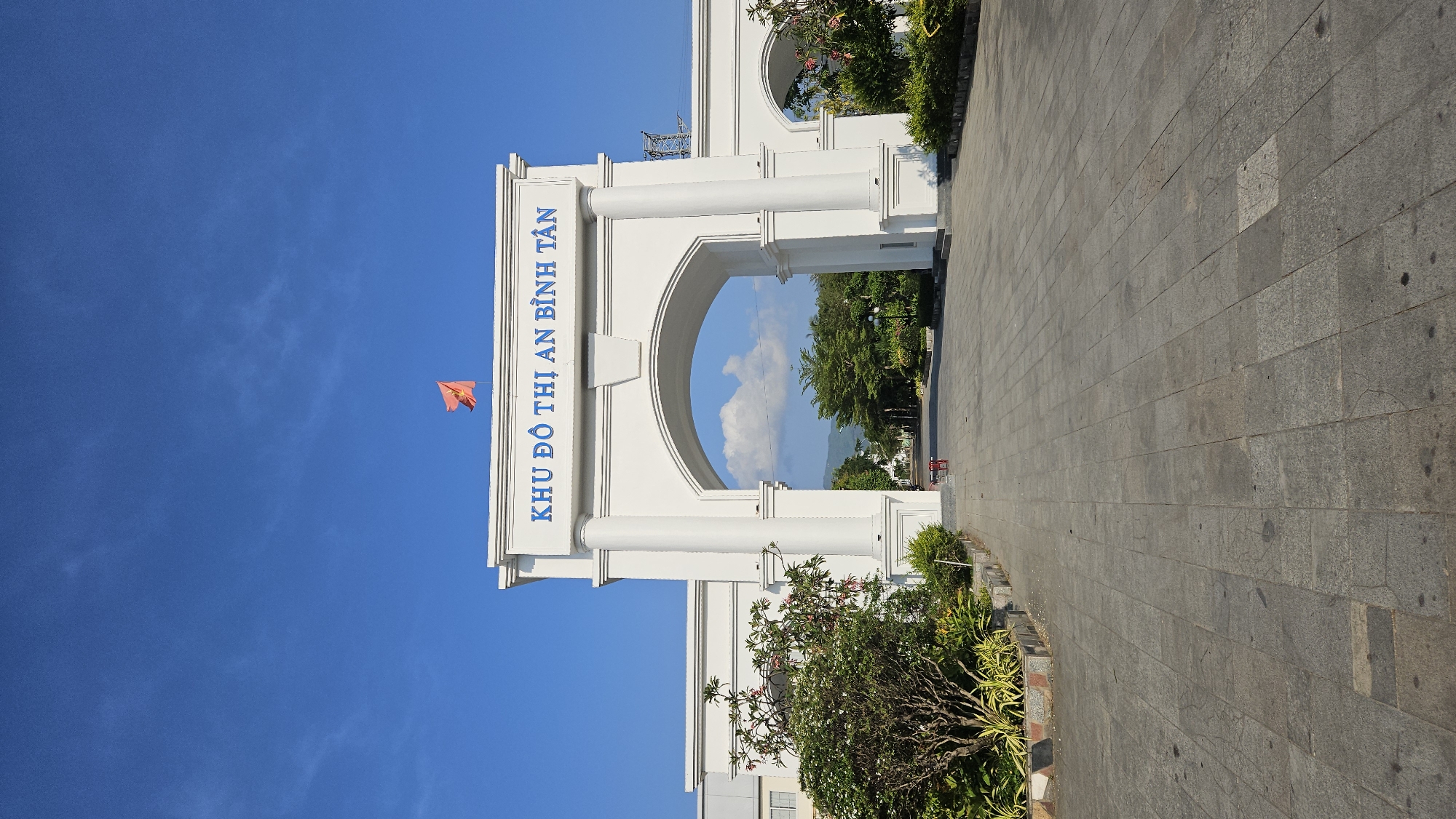 The image shows the entrance gate to the An Binh Tan residential area in Nam Nha Trang, Vietnam. The gate is a large, white, arched structure with blue lettering. A Vietnamese flag is mounted on the gate. Through the archway, trees, a street, and distant mountains are visible. The foreground is paved with gray stones. The sky is blue with some clouds. Shrubs and flowers are planted near the gate.