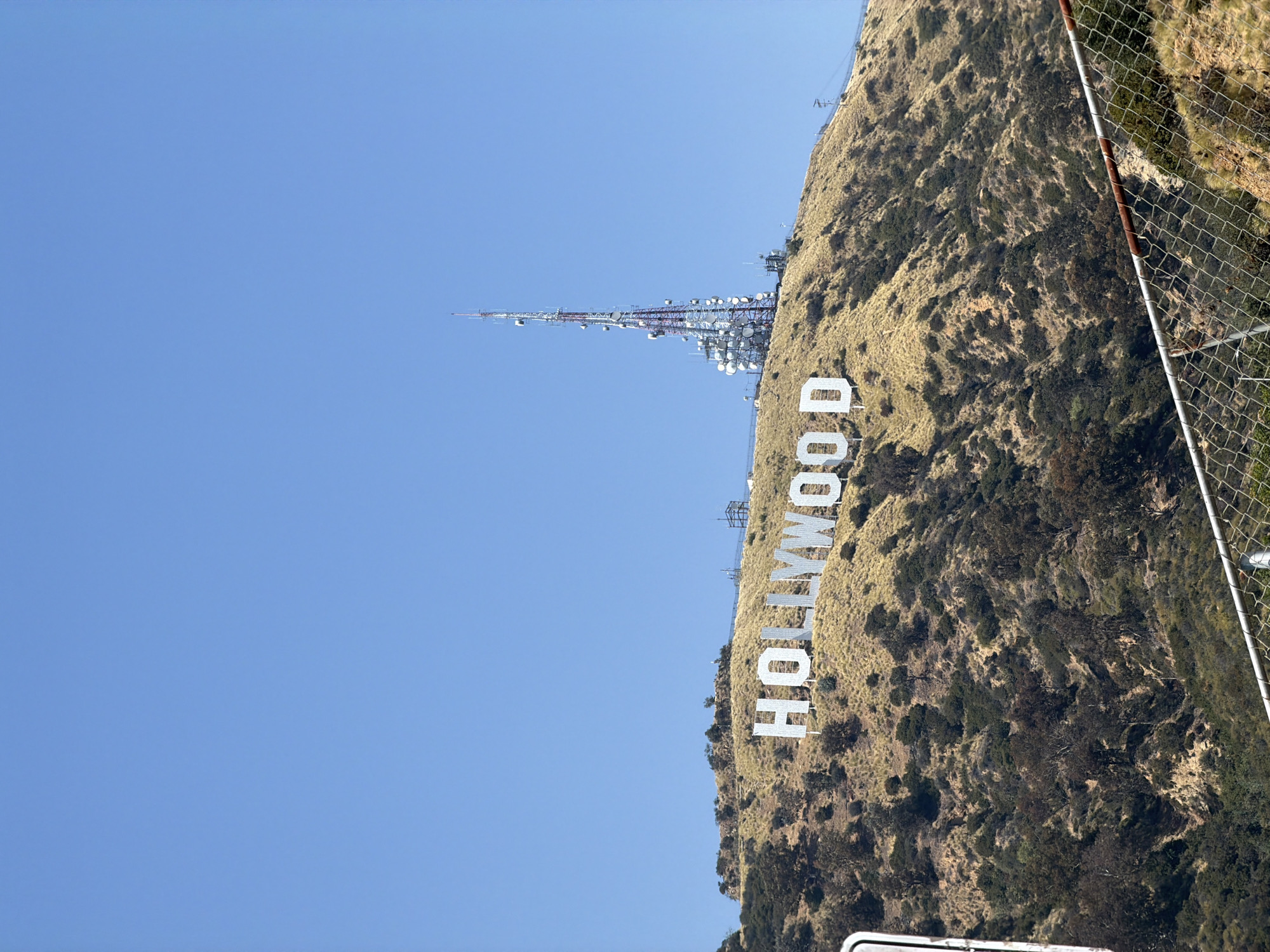 The image depicts the Hollywood Sign, a famous landmark in Los Angeles, California. The sign is positioned on a hillside covered with vegetation. Above the sign, a tall communication tower structure stands on the same hilltop. The sky is clear and blue. A chain-link fence is visible on the right side of the frame.