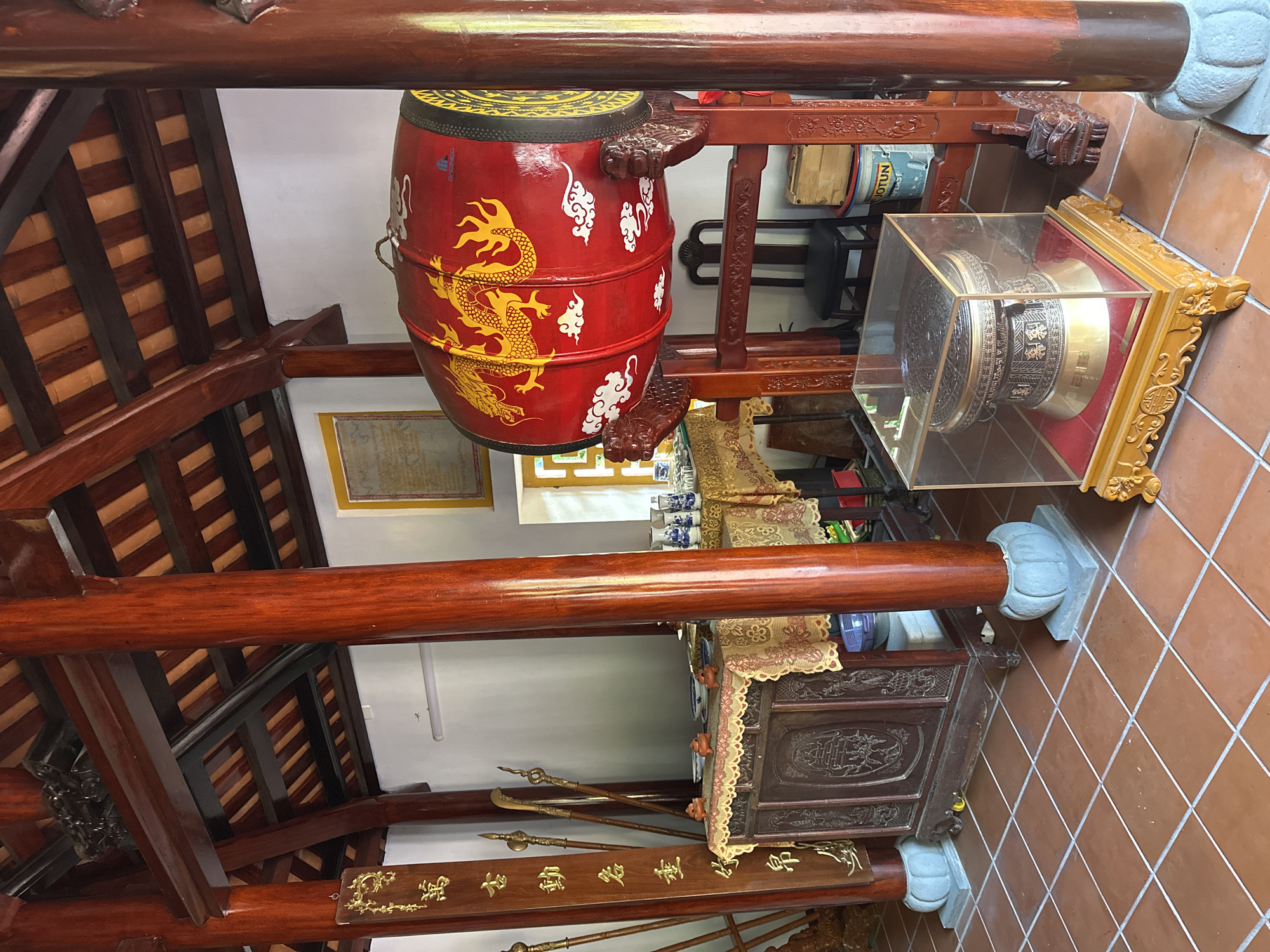 The image depicts the interior of a traditional building, likely a temple or ancestral house, in Da Nang, Vietnam. The structure features prominent red wooden pillars and beams supporting a tiled roof. On the left, a tall wooden plaque inscribed with golden Vietnamese characters stands next to a collection of ornate spears. In the center, a dark wooden altar table is draped with a patterned cloth, displaying small ceramic figures and vases. To the right, a large, red ceremonial drum adorned with yellow dragons and clouds is mounted on a wooden stand. In the foreground, a smaller, intricately decorated drum is showcased within a glass case, resting on a carved wooden base. The floor is tiled with reddish-brown squares. The overall scene conveys a sense of cultural heritage and religious significance.