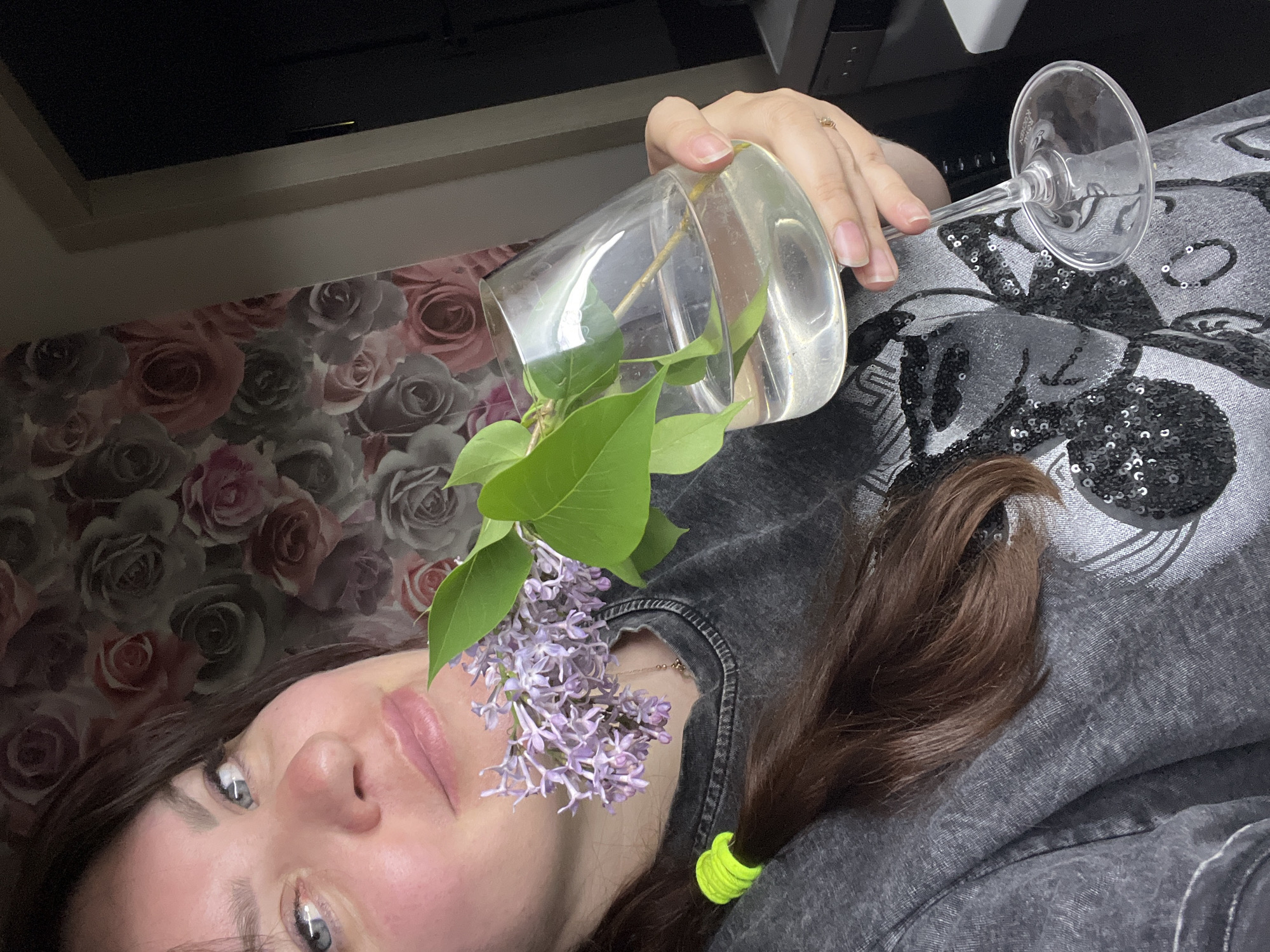 A woman is lying on a gray garment with sequins. In her right hand, she holds a clear glass with water and a lilac flower branch. Part of her hair is tied with a yellow band. The background is a wall with a floral pattern of roses. Behind her head and the glass is a black object. The location is unknown.