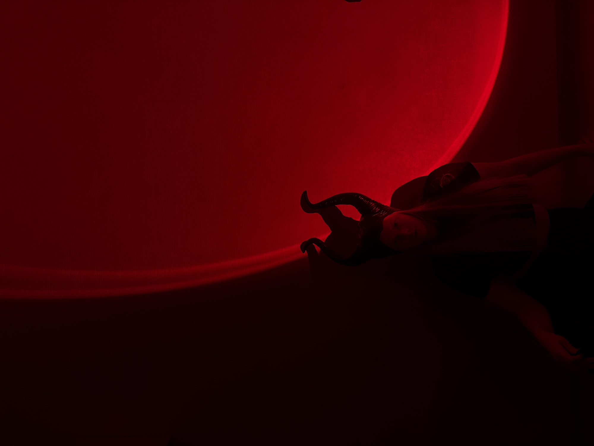This image captures a dramatic and atmospheric scene, dominated by deep red and black tones. A person, likely a woman, is positioned diagonally across the right side of the frame, appearing to be reclining or lying down. She is adorned with a headpiece featuring prominent, dark, curved horns, suggesting a costume or character such as a devil or a mythical creature. Her long, dark hair cascades around her head, blending into the dark surroundings, and she wears dark clothing.
A large, circular projection of intense red light illuminates a textured dark red wall or surface, occupying the upper left and central areas of the composition. A thinner, elongated streak of red light extends from the bottom left corner, adding to the dynamic lighting. The overall lighting is artificial, low-key, and creates significant areas of deep shadow, contributing to a mysterious and intense mood. The person's face, partially visible, is softly illuminated with a red glow, displaying a neutral expression. The setting is likely an indoor space in Izhevsk, Russia, used for an artistic or thematic photoshoot. No text is visible in the media file.