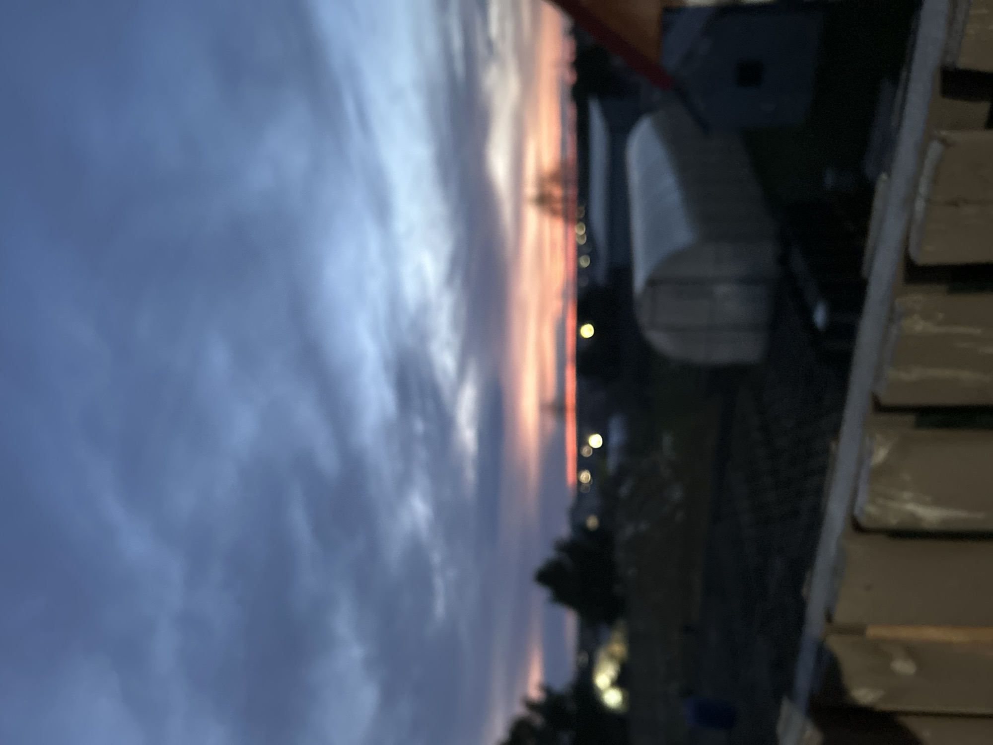 This blurry image, taken in Фоки, Russia, captures an outdoor scene at either sunrise or sunset. The sky dominates the upper portion of the frame, displaying a gradient of colors from a dark, cloudy blue-grey at the top to warm, soft hues of peach, orange, and pink along the horizon. Diffused clouds are visible across the sky.
Along the distant horizon, a scattering of small, circular bokeh lights suggests the presence of a town or city, along with indistinct dark silhouettes of trees or buildings. In the midground, to the right, a light-colored, possibly semi-cylindrical structure, such as a greenhouse or a roof, is partially visible. The immediate foreground on the right side of the image features a structure composed of stacked horizontal planks, possibly a fence, wall, or balcony railing, cast in partial shadow.
The entire image is significantly out of focus, creating a soft, dreamlike quality. No people are visible, and no specific activities are taking place. There is no visible text. The overall impression is one of serene atmospheric conditions, typical of twilight hours.