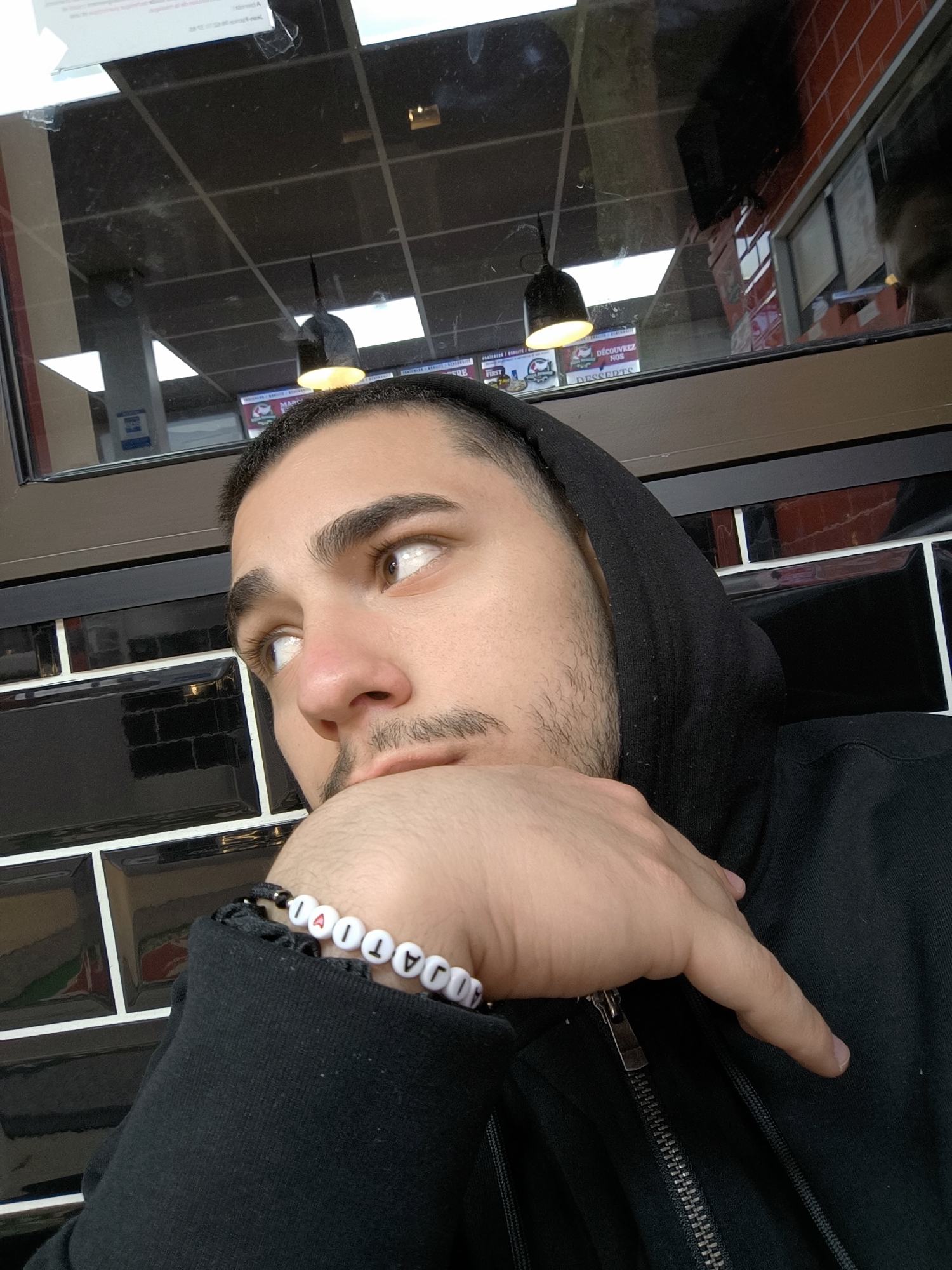A medium shot depicts a light-skinned male with brown hair and stubble wearing a black hooded sweatshirt. He is resting his chin on his right hand, and a bracelet of white beads with black lettering on it is visible on his wrist. The beads spell out “4IJAT1VAL”. He is looking slightly upward and to the left with brown eyes. The background consists of black tile with white grout, and above the tile is a window reflecting ceiling lights and a red tiled area. The location is in Strasbourg, France, as indicated by a sign visible in the window reflection stating "Découvrez nos DESSERTS".