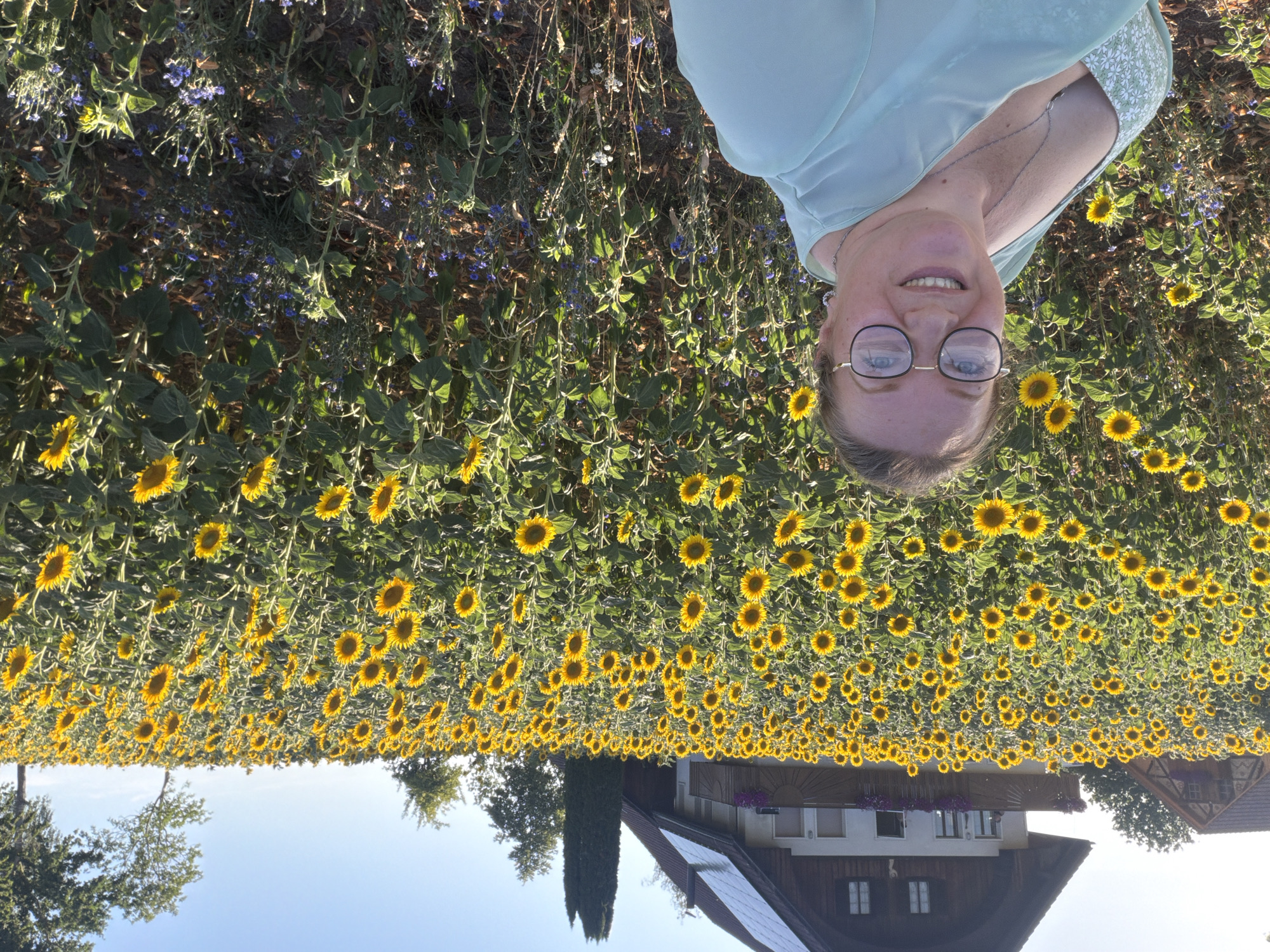 A woman wearing glasses and a pale green dress smiles at the camera in front of a vast field of sunflowers. The field extends into the background, with tall sunflowers dominating the scene under a clear blue sky. In the distance, a traditional Swiss-style house with solar panels on its roof is visible. The woman is positioned in the foreground, on the left side of the image, with the sunflowers filling the majority of the frame behind her. Small blue wildflowers are interspersed among the sunflowers. The scene is set in Ufhusen, Switzerland, bathed in natural sunlight, suggesting a warm day.