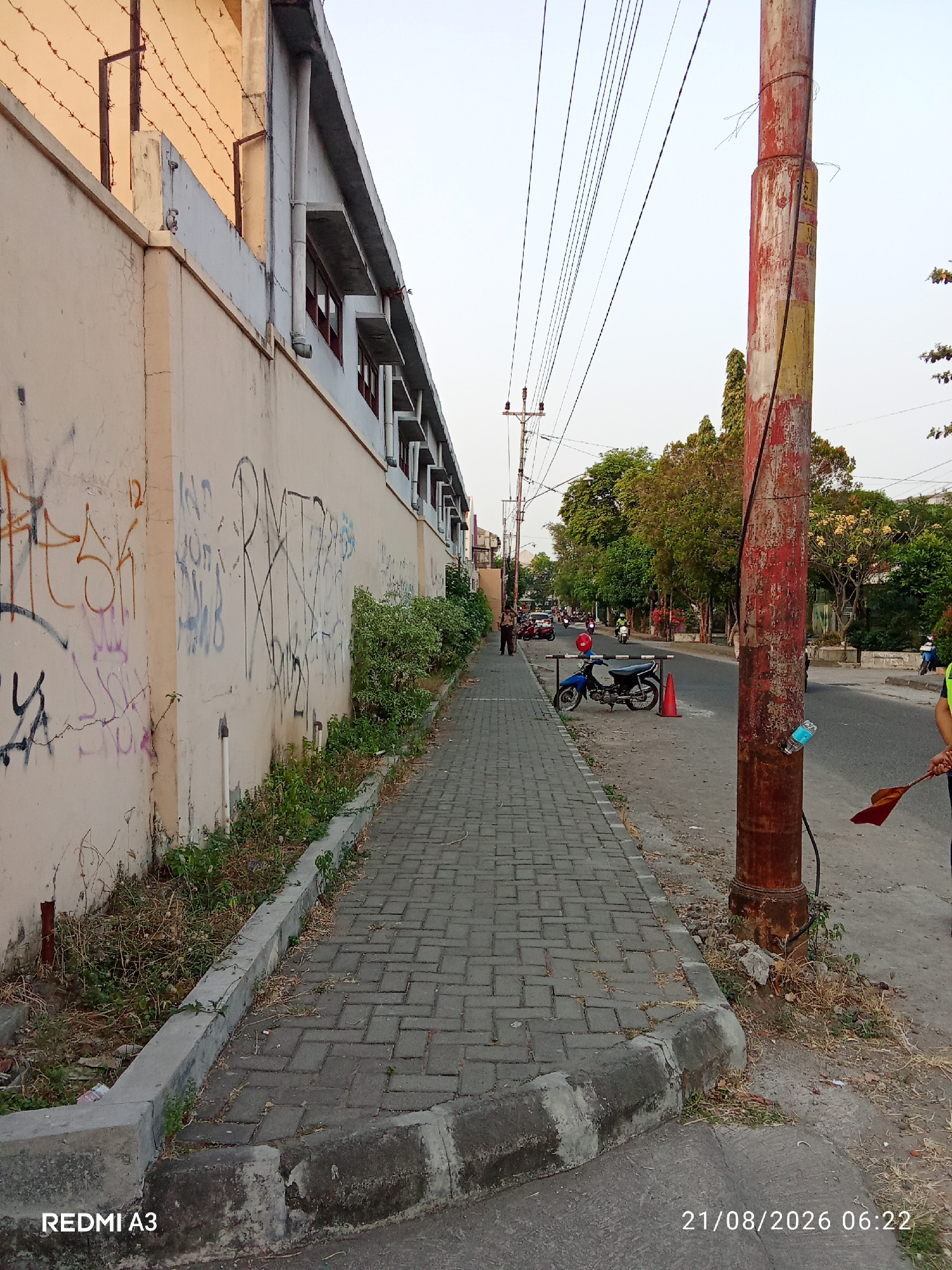 The image depicts a sidewalk alongside a wall covered in graffiti, with a street running parallel to it. In the background, trees and buildings are visible. The scene appears to be in an urban environment, possibly in Surakarta, Indonesia, given the timestamp on the image.
The sidewalk is paved with gray bricks, and a concrete curb separates it from the street. Along the wall, there is a strip of overgrown weeds and dry grass. The wall itself is painted a light beige color and is heavily marked with various graffiti tags in black, red, and purple. The building adjacent to the wall has a concrete structure and several windows with red frames.
On the street, several motorcycles are parked or being ridden. A blue motorcycle with a red ball on its handlebars is parked near an orange traffic cone. A utility pole, painted red and yellow, stands on the right side of the image, with electrical wires extending from it. A plastic bottle is attached to the pole. A person in a yellow vest is visible on the far right, holding a red flag. The sky is clear and overcast, suggesting it might be morning or late afternoon. The timestamp at the bottom right corner of the image reads "21/08/2026 06:22", indicating the date and time the photo was taken. The text "REDMI A3" is visible in the bottom left corner, likely identifying the camera model used to capture the image.