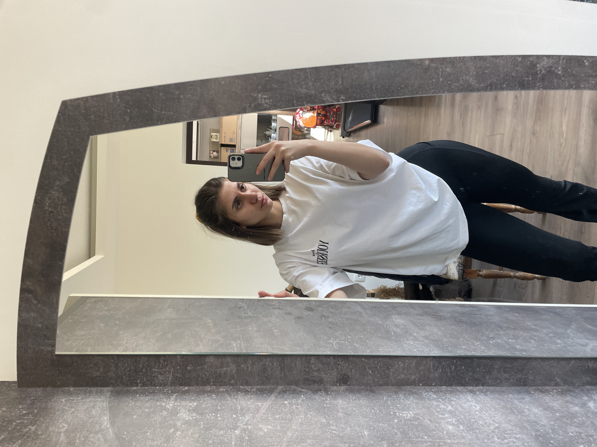 A woman is taking a selfie in a mirror. The mirror has a grey frame with a curved top. The woman is wearing a white shirt and black pants. She is sitting in a chair with wooden legs. The background includes a shelf with objects, a child's stroller, and a wooden floor. The location is Sofia, Bulgaria.