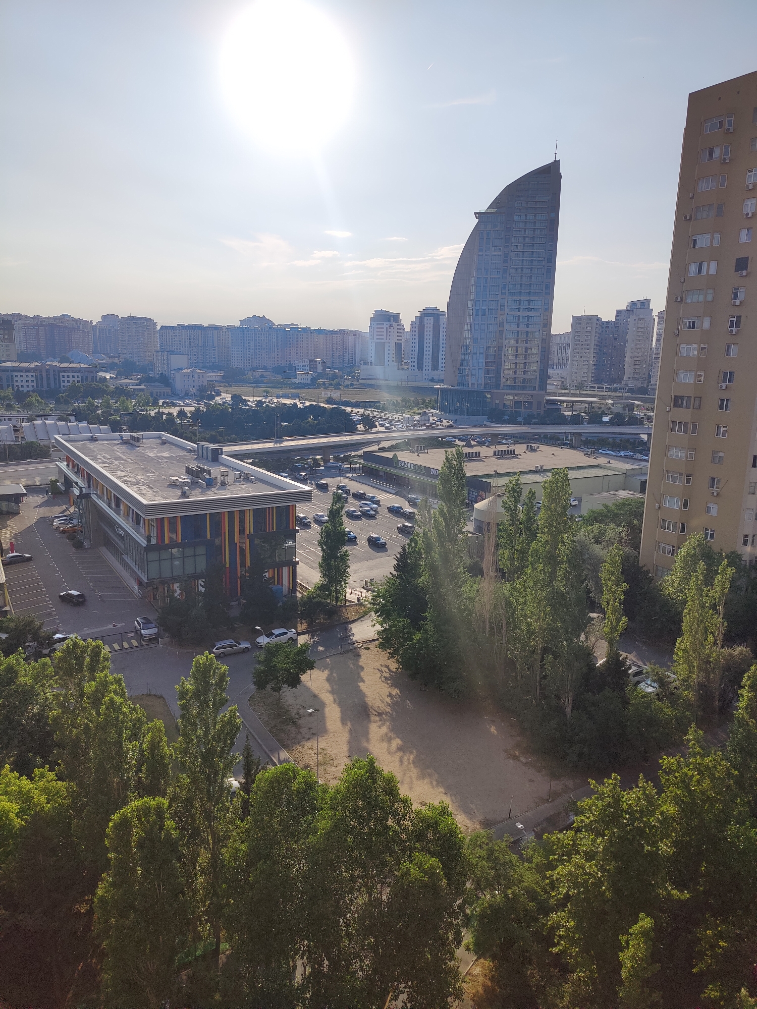 The image depicts an aerial view of a cityscape, likely in Baku, Azerbaijan, under a bright, sunny sky with lens flare. In the foreground, a dense canopy of green trees dominates the lower portion of the frame. Beyond the trees, a modern, multi-story building with a colorful facade, featuring vertical stripes of red, yellow, and blue, is visible. This building has a flat roof with visible equipment. A parking lot with several cars is situated in front of this building.
Further into the midground, a curved, glass-fronted skyscraper with a distinctive architectural design stands tall against the backdrop of numerous other residential and commercial buildings that form the city skyline. Another tall, beige apartment building with many windows and air conditioning units is positioned to the right. A multi-lane road with moving vehicles is present, suggesting urban activity. In the distance, the urban landscape stretches out, characterized by a multitude of buildings of varying heights and styles. The overall impression is one of a bustling, modern city bathed in sunlight.