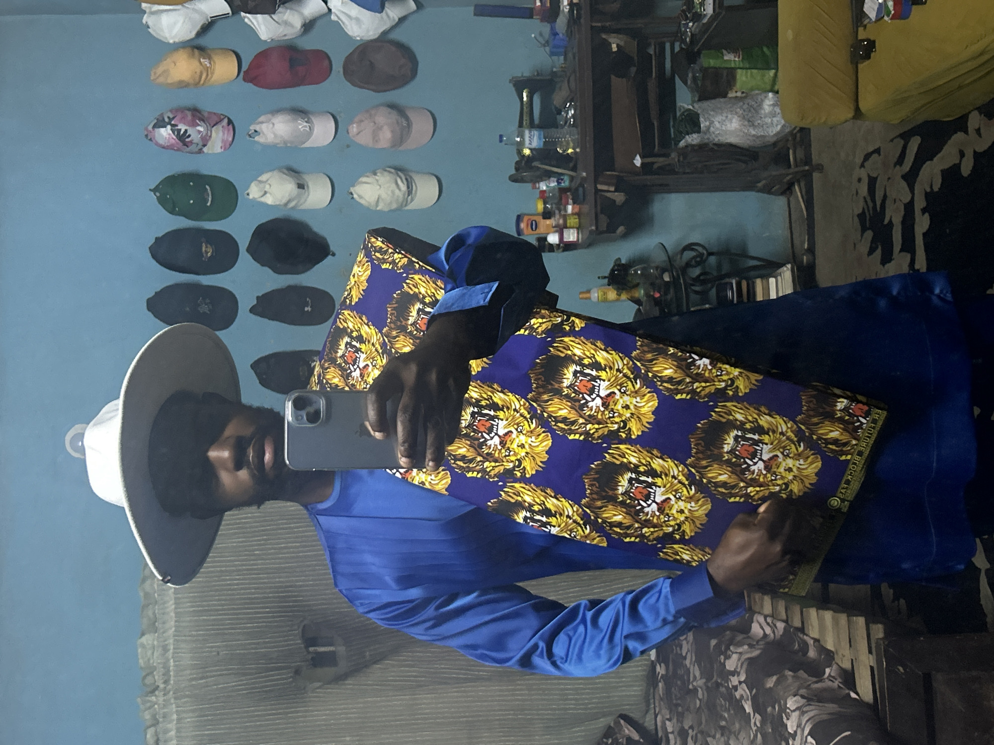 A young Black man, wearing a white wide-brimmed hat and a bright blue, long-sleeved garment, stands in what appears to be a workshop or tailor's studio in Lagos, Nigeria. He is holding a piece of fabric printed with a vibrant pattern of lion faces on a deep purple background. The man is taking a selfie with a smartphone. In the background, a wall is adorned with numerous baseball caps and other headwear. To the right, a vintage sewing machine sits on a wooden table cluttered with various items, including bottles and tools. A yellow sofa is visible in the lower right corner. The overall impression is that of a creative space where textiles and fashion are central.