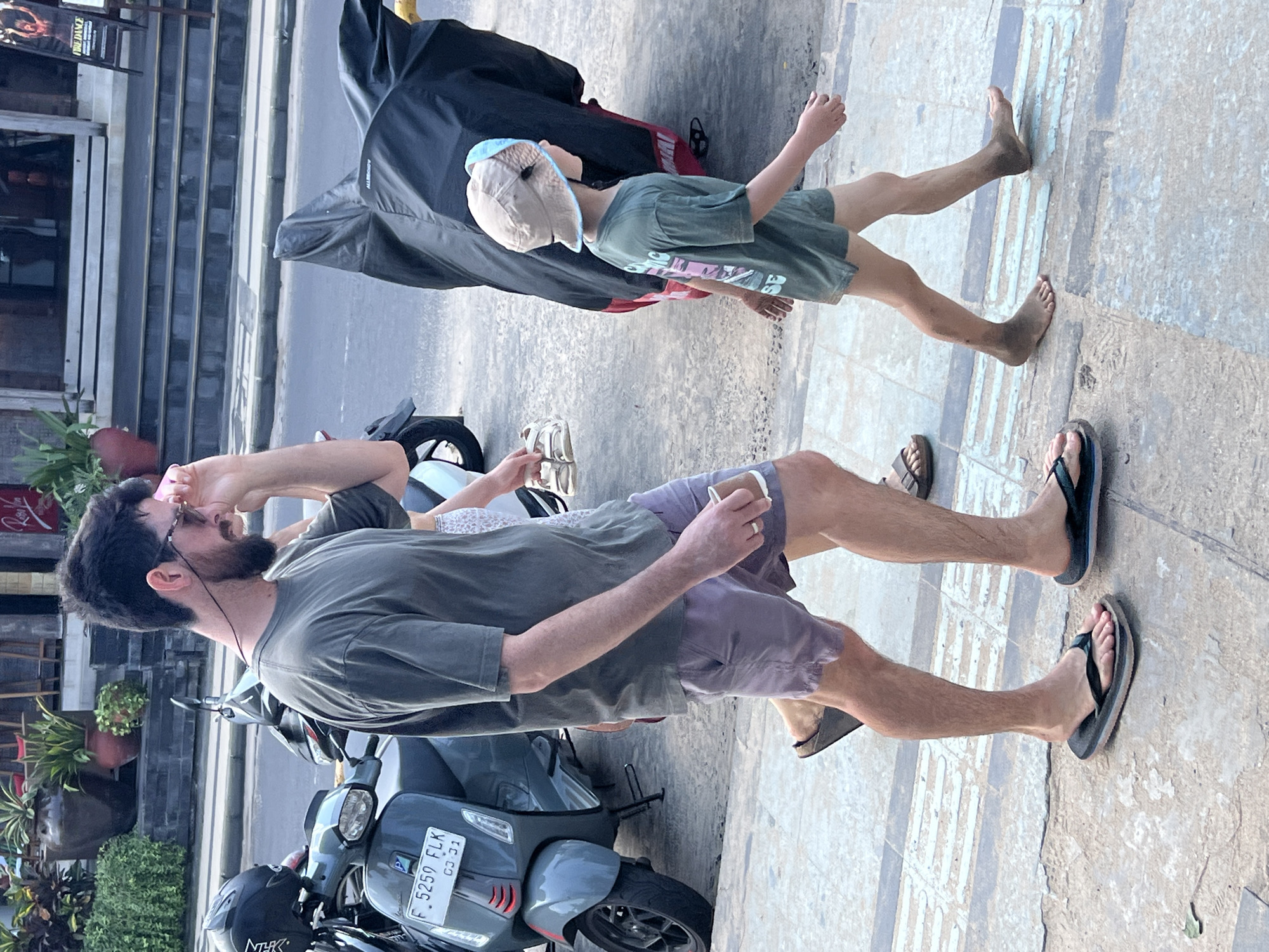 A man and a young boy walk on a concrete sidewalk in Kuta, Indonesia. The man is wearing a green t-shirt and purple shorts, and is holding a small brown cup in his right hand. He is wearing flip-flops on his feet. The boy is wearing a green t-shirt and khaki shorts, and a tan hat with a blue brim. His feet are bare and appear dusty. They are walking away from the camera. To the left of the man is a gray scooter. To the right of the boy is a motorcycle covered with a black tarp. The background shows a street with shops and potted plants.