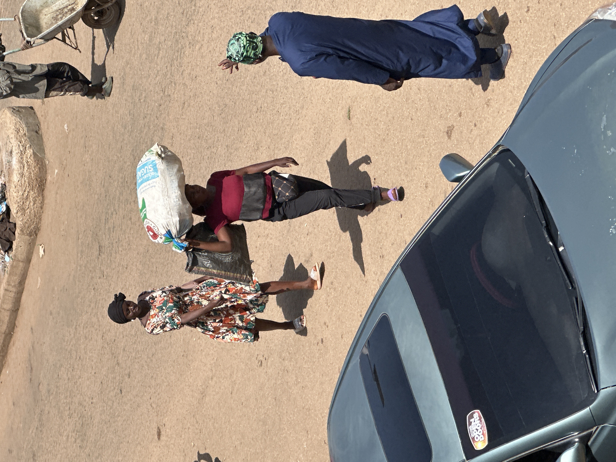 In Bukuru, Nigeria, a scene unfolds on a dusty, sun-drenched street. Three individuals are visible. In the foreground, the hood of a teal sedan with a sunroof is prominent. On the left side of the road, a woman wearing a black headscarf and a colorful floral dress walks, her head tilted slightly. To her right, another woman carries a large white sack of what appears to be sugar on her head. She wears a red and grey striped top, black pants, and a patterned fanny pack. Her left foot is adorned with a purple sandal. Further to the right and slightly behind, a man wearing a blue robe and a green patterned headscarf stands with his right arm raised as if to greet someone or direct traffic. In the background, a person in a grey top and dark pants pushes a wheelbarrow. The environment is characterized by a dry, sandy ground, with some debris and a rough concrete border visible. The lighting suggests a bright, sunny day, casting distinct shadows.