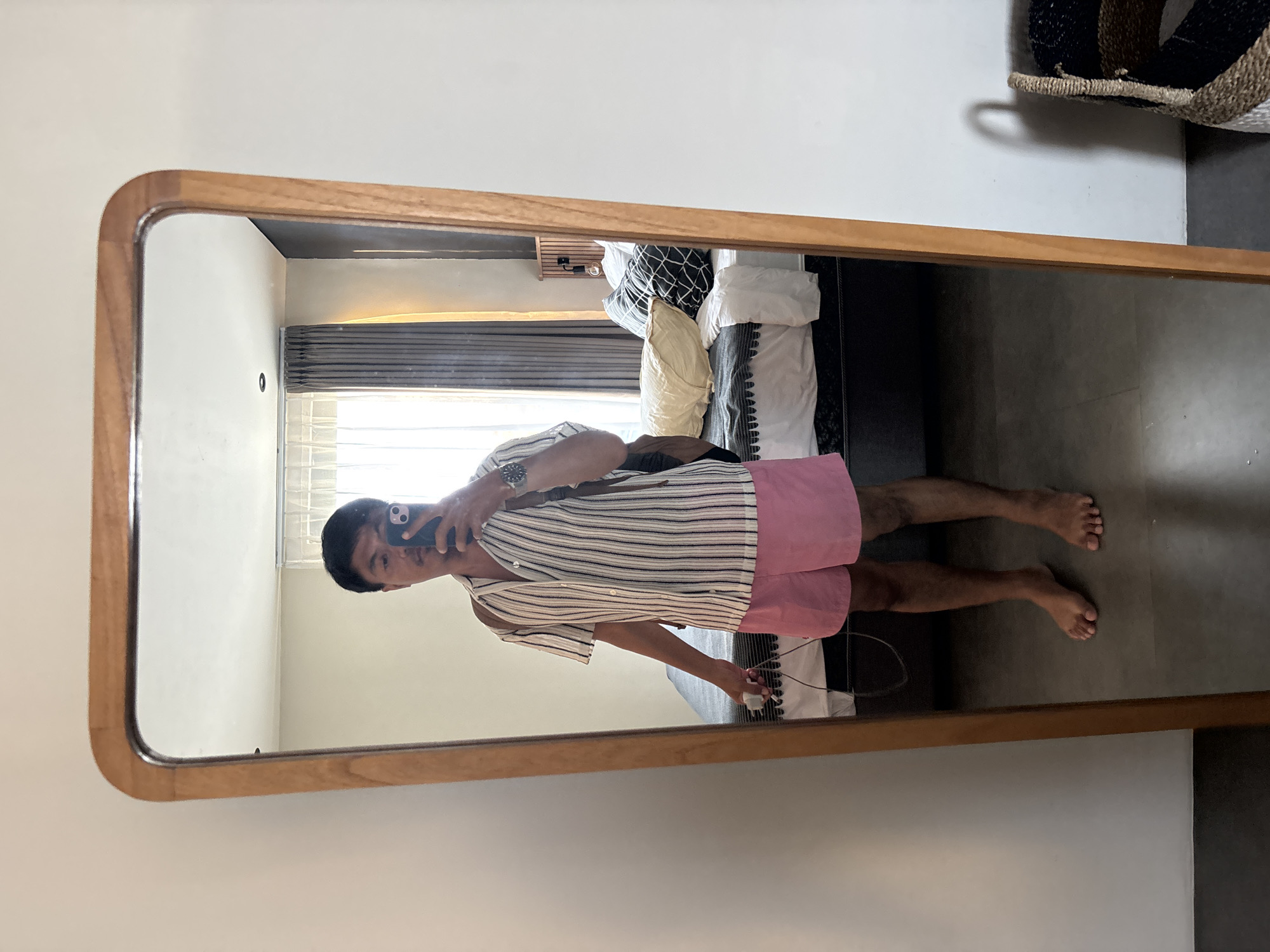 A person is taking a mirror selfie in what appears to be a bedroom. The person is wearing a vertically striped short-sleeved button-down shirt and pink shorts. They are holding a smartphone in their right hand and a white object with a cord in their left. A brown backpack is slung over their right shoulder. In the mirror's reflection, a bed with pillows and a headboard is visible. The room has white walls and a light-colored floor. The mirror itself has a rounded wooden frame. The scene is located in Mengwi, Indonesia.
