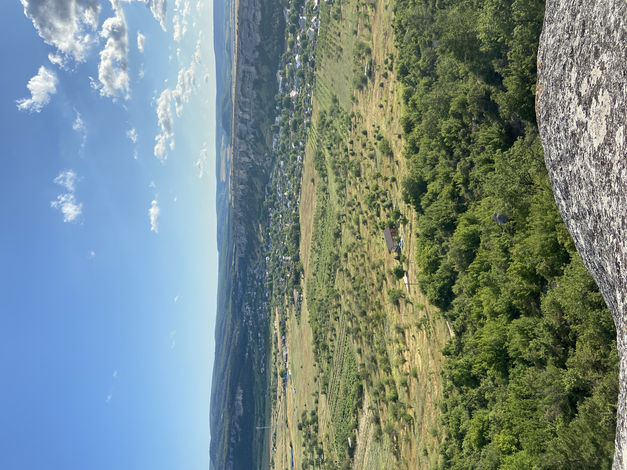 This is a high-angle, full shot of a rural landscape under a bright blue sky with scattered white clouds. In the foreground, a large, grey, textured rock juts into the frame from the bottom. Beyond the rock, a dense area of lush green trees forms a verdant canopy. This transitions into a more open, undulating valley dotted with cultivated fields, some appearing golden and dry, others greener and showing rows of young trees. A small village is visible in the middle ground, nestled amongst the greenery, with clusters of houses, some with red roofs. In the far distance, a line of imposing, sheer, light-colored cliffs rises dramatically, forming a natural barrier. The overall impression is one of a serene and expansive rural scene, likely taken from a viewpoint overlooking the village and the surrounding countryside.