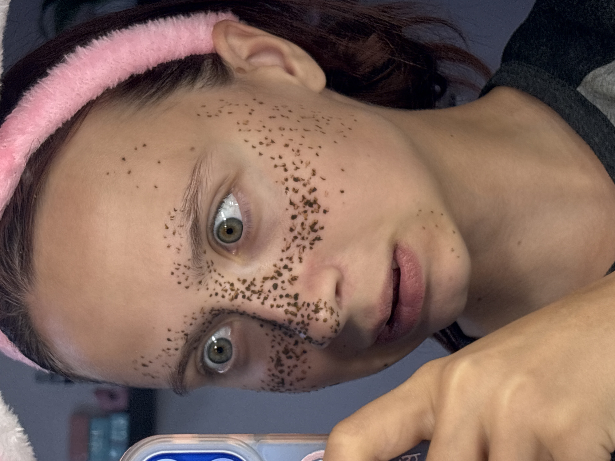 A close-up shot features a young woman with green eyes and light skin, adorned with an artificial dusting of freckles across her face, including her nose and cheeks. She is wearing a pink fuzzy headband and holding up a phone with her left hand, her mouth slightly agape in an expression of surprise or wonder. The background is dimly lit, with a hint of shelves and books visible on the left, suggesting an indoor setting, possibly a bedroom. The overall lighting is soft, highlighting the woman's face and the details of her makeup. There is no discernible information that can place this image in the city of Прикамский, Russia.