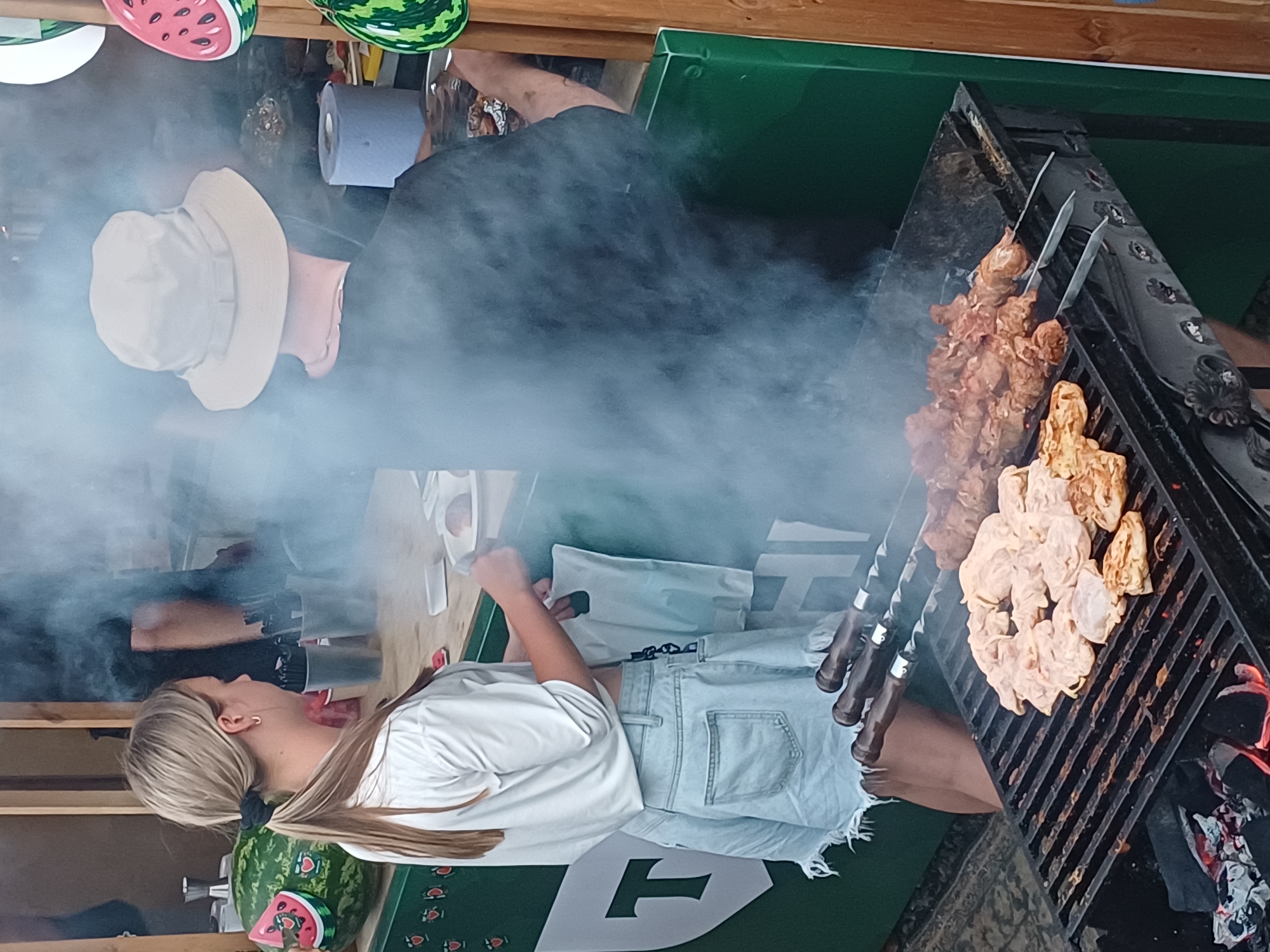 The image depicts an outdoor scene, likely at a street food stall in Estosadok, Russia. In the foreground, a charcoal grill is actively cooking various meats on skewers. The meats appear to be chicken pieces, some marinated and others grilled to a golden brown. Smoke from the grill billows upwards, partially obscuring the figures in the background.
A young woman with blonde hair tied in a ponytail is standing to the left of the grill. She is wearing a white cropped t-shirt and light-wash denim shorts. She is holding a white plastic bag. To her right, a person wearing a light-colored bucket hat is standing, their back to the camera. They are wearing a dark t-shirt and their arms are visible, with tattoos on their forearms. The person is holding what appears to be tongs or another grilling utensil.
Behind the grill, a large watermelon decoration is visible on the left. The stall itself is decorated with green panels and features watermelon motifs. A roll of blue paper is visible on a counter behind the grill. The overall atmosphere suggests a casual, possibly festive, outdoor dining or market environment.