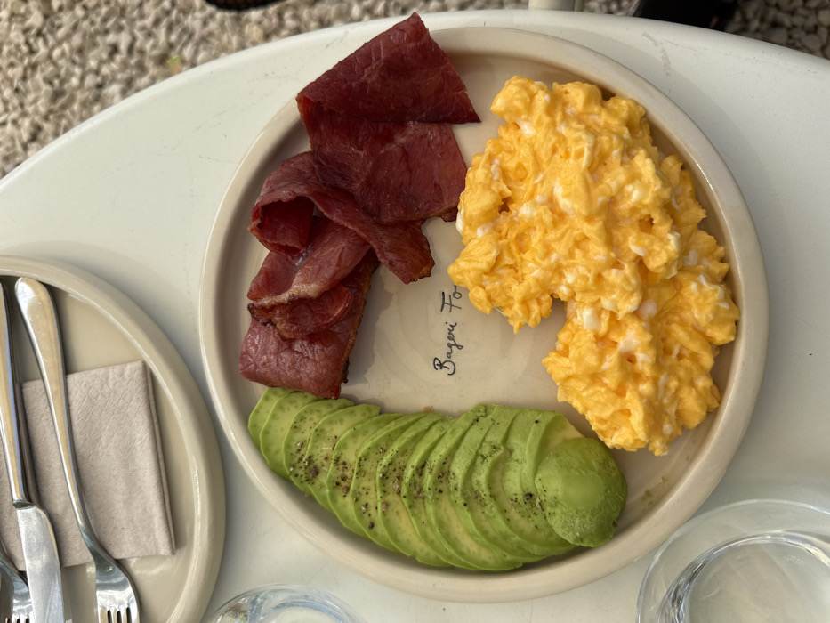 This is an overhead shot of a breakfast plate served in Dubai, United Arab Emirates. The primary subject is an oval, cream-colored ceramic plate displaying a meal composed of three distinct items. On the left, approximately ten slices of fresh green avocado, seasoned with black pepper, are arranged linearly. In the center, several cooked, reddish-brown strips of meat, consistent with beef bacon or thinly sliced cured meat, are stacked. Below the meat and to the right, a generous portion of bright yellow scrambled eggs occupies the plate. Faint black script reading 'Bageri Fo' is visible on the plate surface. The plate rests on a white table. To the upper left, a second, identical cream-colored plate is partially visible, holding a silver-colored fork and knife neatly placed on a beige napkin. A clear glass, partially filled with water, is visible on the table to the lower left. The background to the far right shows a blurred textured surface, likely outdoor ground covering. The scene is brightly lit, suggesting daylight.