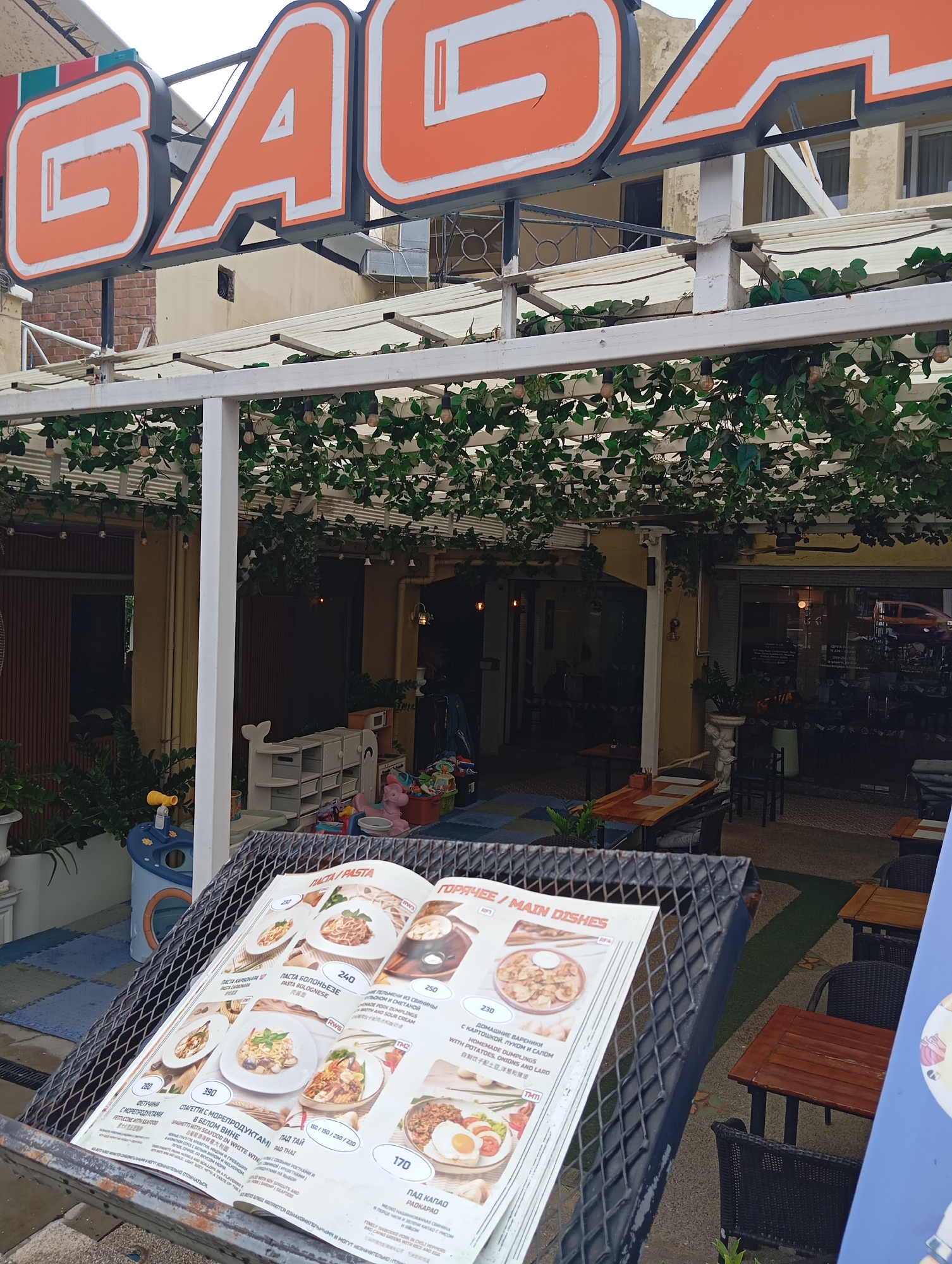 A casual restaurant is depicted in the city of Karon, Thailand. The restaurant features a bright orange and white sign with the word "GAGA" in large letters. The outdoor seating area is decorated with artificial greenery and string lights, creating a relaxed ambiance. A menu is displayed on a metal stand in the foreground, showcasing various food items. Some of the dishes are identified by name in both English and what appears to be Russian. The menu also includes prices in what could be local currency, indicated by numbers like 240, 250, and 170. Beyond the seating area, the entrance to the restaurant is visible, with glass doors and windows revealing an indoor space. The overall impression is of a welcoming establishment offering a diverse menu in a pleasant setting.