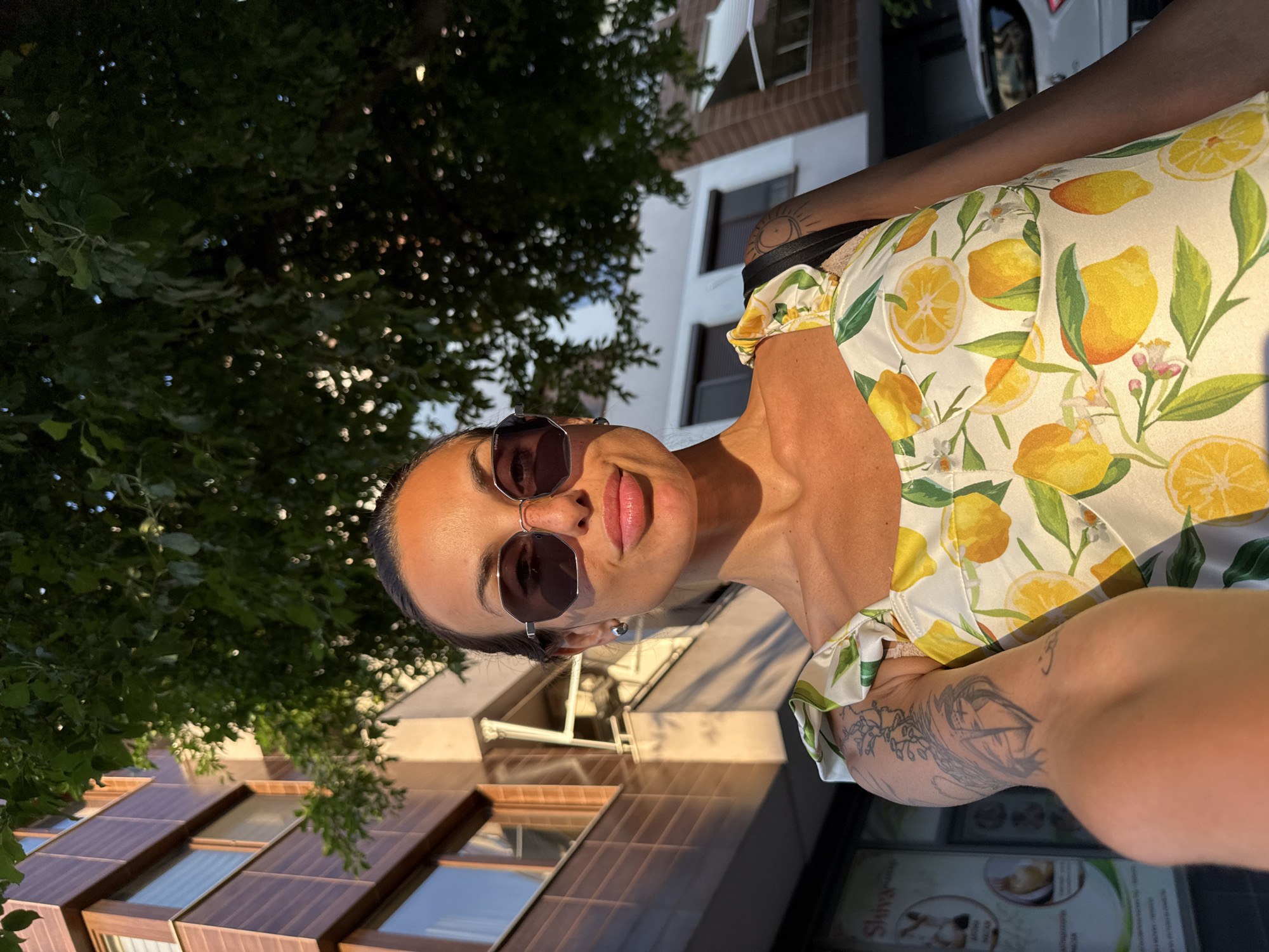 A woman with sunglasses and a lemon-print dress is captured in a selfie. The image is taken outdoors with a building and trees in the background, suggesting an urban or suburban setting. Sunlight illuminates the woman's face and upper body, indicating it's likely daytime, possibly late afternoon due to the warm lighting. She has tattoos visible on her arm. The building in the background has multiple windows with a reflective surface, possibly glass or metal. Part of a car is visible to the right. The image appears to be taken in Pleven, Bulgaria. The woman's expression seems relaxed and cheerful. There is text visible on a sign in the lower right corner which reads "Slava".