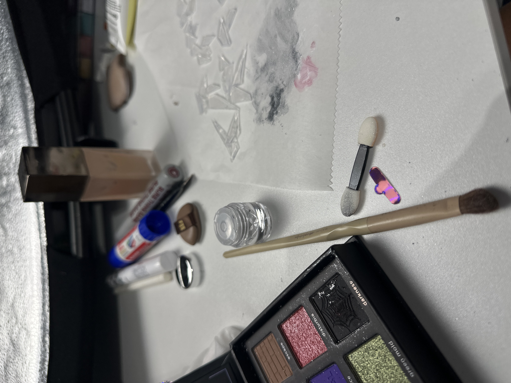The image displays a collection of makeup and art supplies arranged on a white surface, possibly a desk or vanity. In the foreground to the left is an open eyeshadow palette with several shades, including "escape," "disturbia," "darkness," and "green mold." Next to the palette is a small, clear, faceted jar, likely containing cosmetic product or glitter, and a makeup brush with a light-colored handle and a dark, fluffy tip. To the right, a small dual-ended applicator with white sponge tips is visible, along with a few iridescent, shard-like decorative elements. Further back, a tube of glue stick with a blue and red label is present, along with a white cylindrical object that appears to be a makeup pencil or applicator. A tall, rectangular bottle of foundation with a gold cap stands prominently in the upper right quadrant of the image. A piece of white paper with scattered glitter and pinkish smudges is also visible on the surface, along with a few clear, angular pieces that could be nail art embellishments or crystal shards. The background shows a blurred, reflective surface, possibly a light fixture or a mirror. The overall scene suggests a preparation or application process for makeup or a craft project. The image does not provide any specific geographical markers to determine its location in North Kuta, Indonesia.