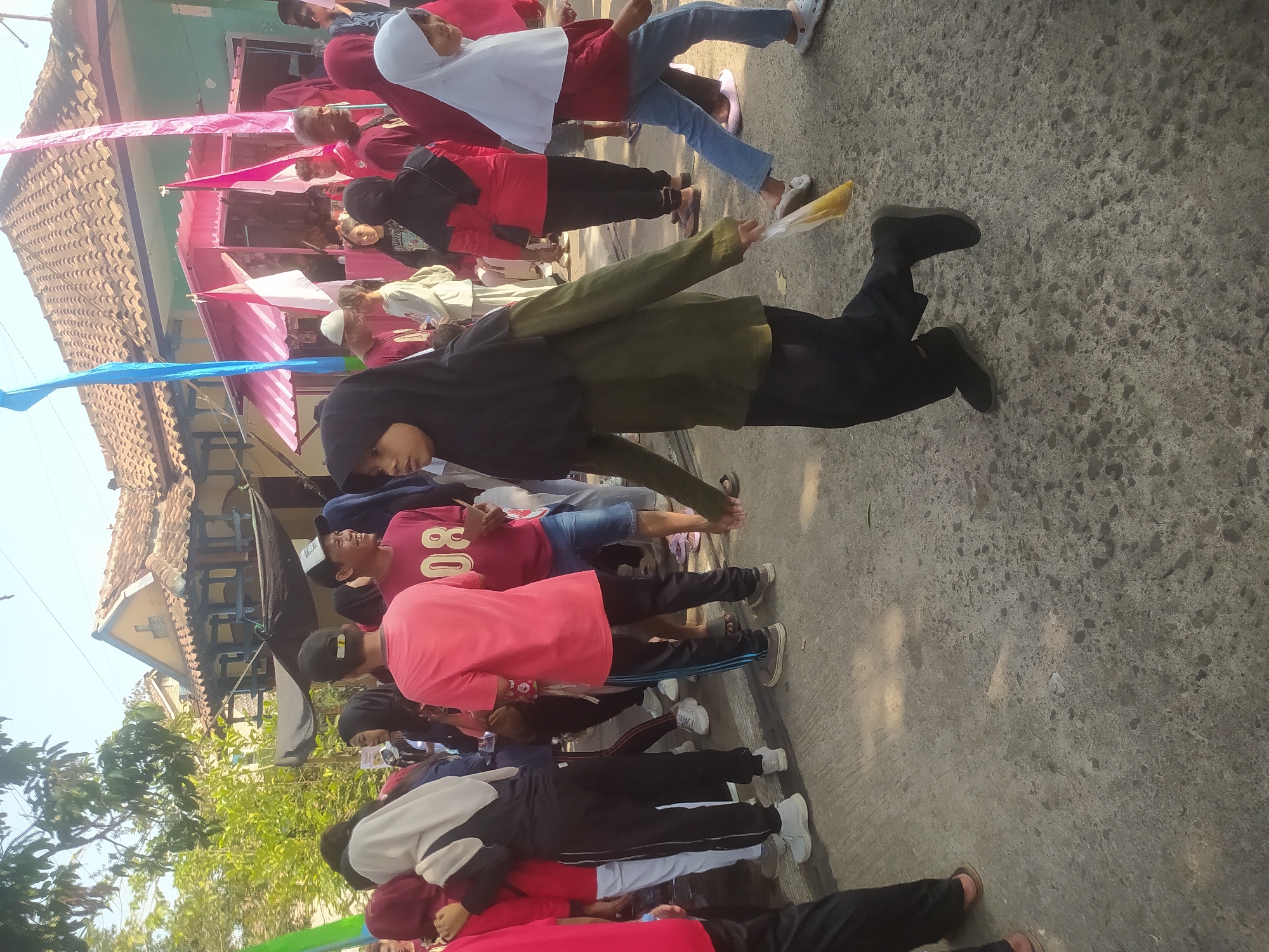 A group of people are gathered on a street, possibly for a local event or celebration, in Lopait, Indonesia. The setting appears to be a village street with a building in the background, featuring traditional tiled roofing. Several individuals, including children and adults, are wearing casual clothing, with some women wearing hijabs. A few people are in the foreground, and the overall atmosphere suggests a daytime gathering. The ground is paved with concrete, and there are some trees and foliage visible. Decorative banners, possibly flags, are also present, adding to the festive ambiance.