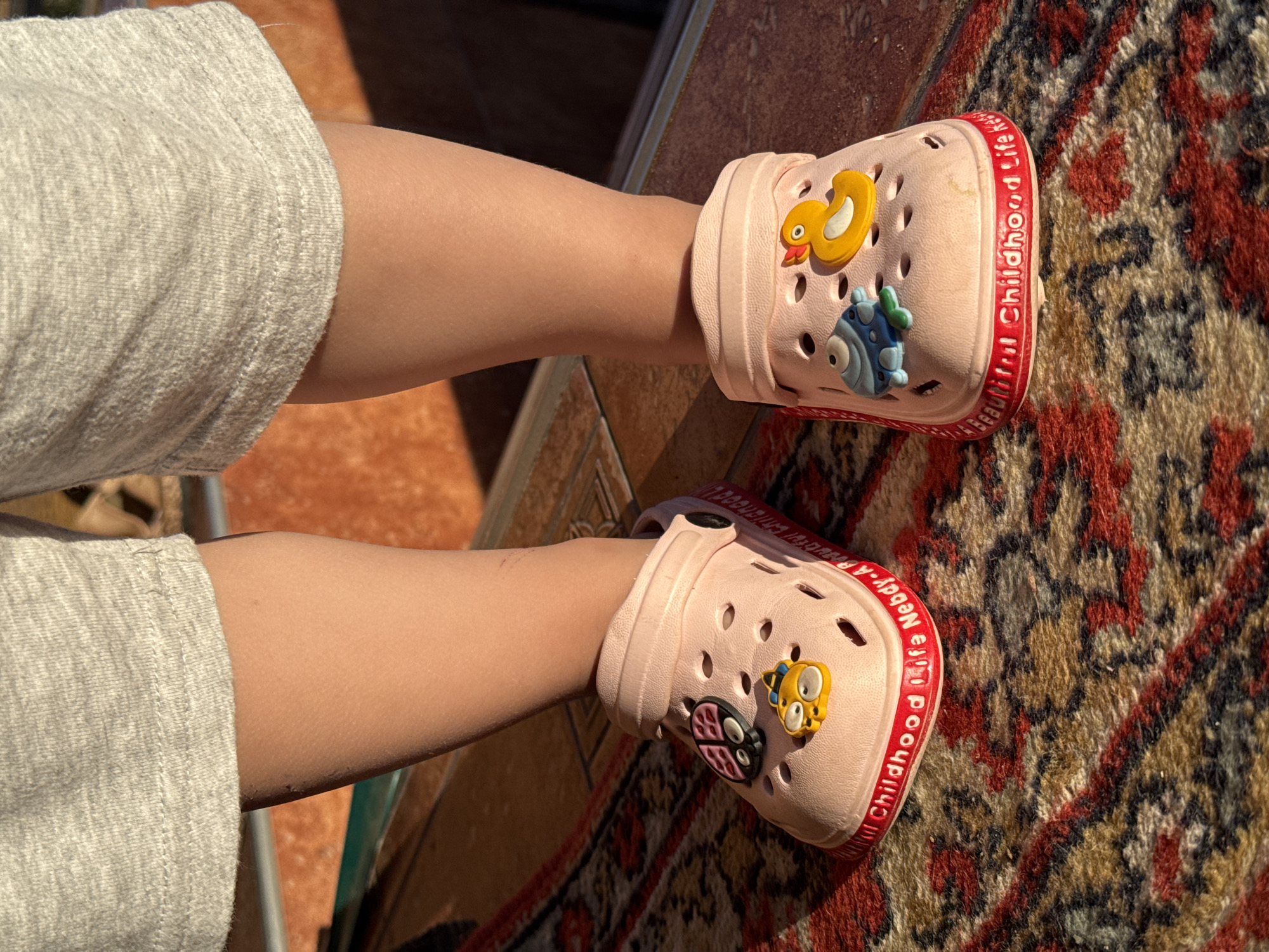 The image shows a close-up view of a child's legs wearing pink clogs adorned with cartoon charms of a duck, a fish, and a ladybug. The clogs have the words "Beautiful Childhood Life" and "Neby.A" printed in red around the sole. The child is wearing grey shorts. The scene appears to be outdoors, with sunlight casting shadows and highlighting the texture of a patterned rug beneath the child's feet. The rug has a complex design with dominant colors of red, brown, and black. The overall impression is one of a warm, sunny day, likely outdoors, capturing a moment of childhood play or relaxation. The setting appears to be a patio or outdoor area, given the tiled surface visible in the background and the rug. The focus is on the child's footwear and legs, suggesting an emphasis on the comfort and fun associated with these shoes. The lighting suggests it is daytime, possibly late morning or afternoon, as the sunlight is strong and creates distinct shadows. The location context provided is Minusinsk, Russia.