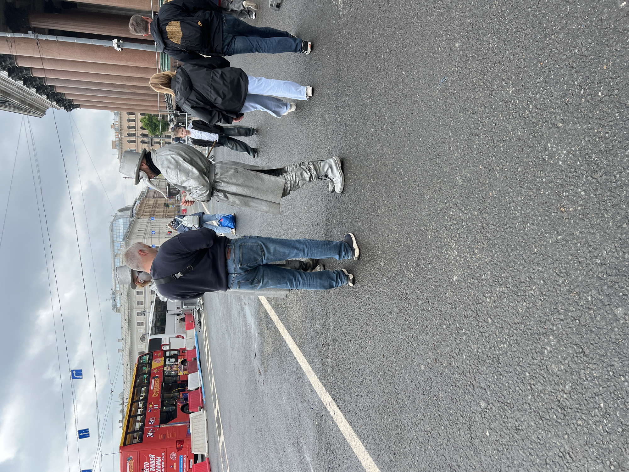 The image depicts a street scene in Saint Petersburg, Russia, near the Winter Palace. A red double-decker tour bus is parked on the side of the street. Several people are walking and standing on the pavement, including a person dressed in silver costume, including a hat and coat. Other people are wearing casual attire such as jeans and jackets. The street surface is asphalt, marked with white lines. The sky is overcast. Buildings with classical architectural features and columns are visible in the background.