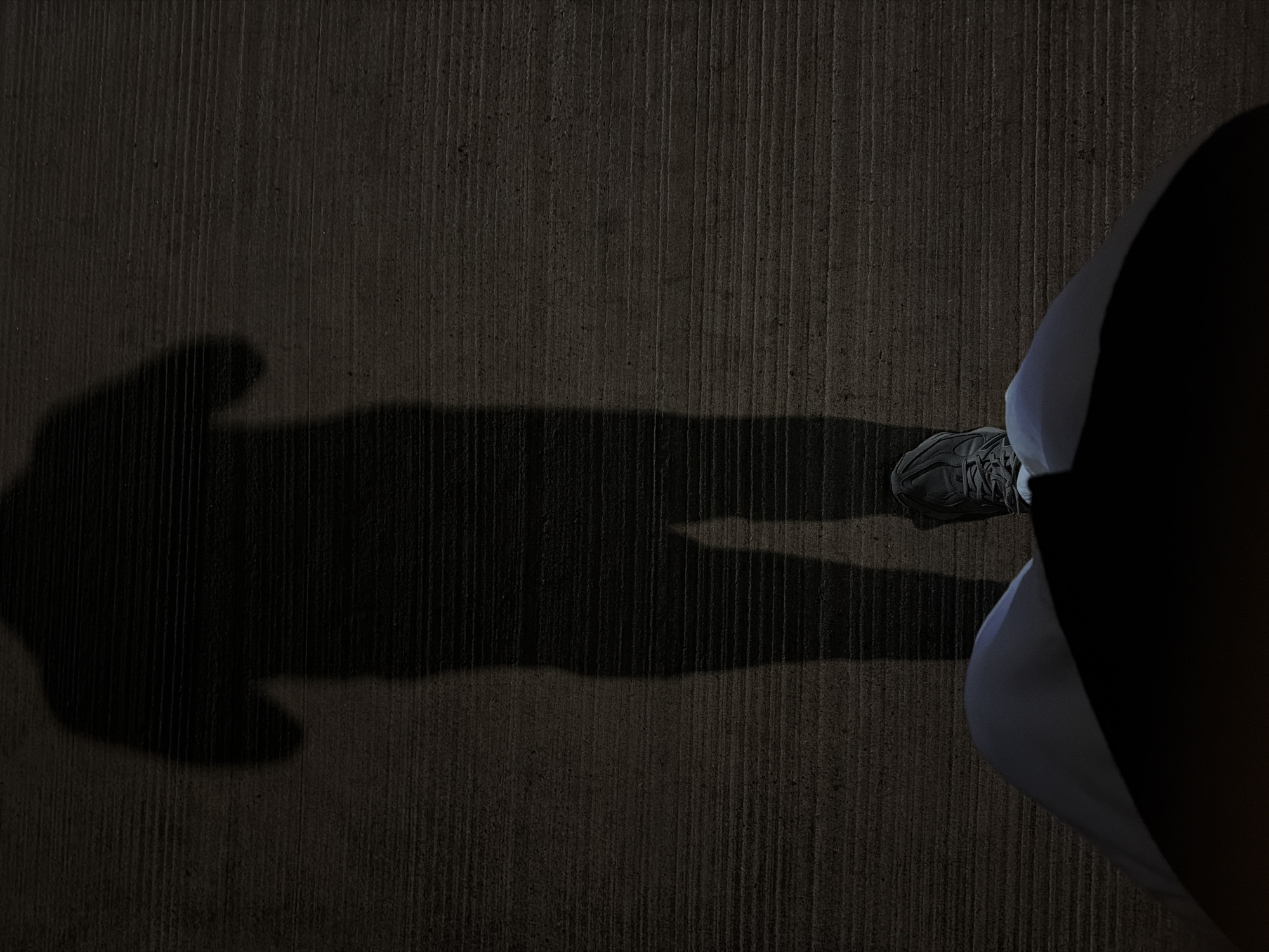 The image shows a person standing on a textured concrete surface, likely a sidewalk or walkway, at night. A long, distorted shadow of the person stretches out to the left. The person is wearing sneakers and light-colored pants. The only part of the person visible is their leg and shoe. The environment is dark. There is no indication of the location being in Fürth, Germany.