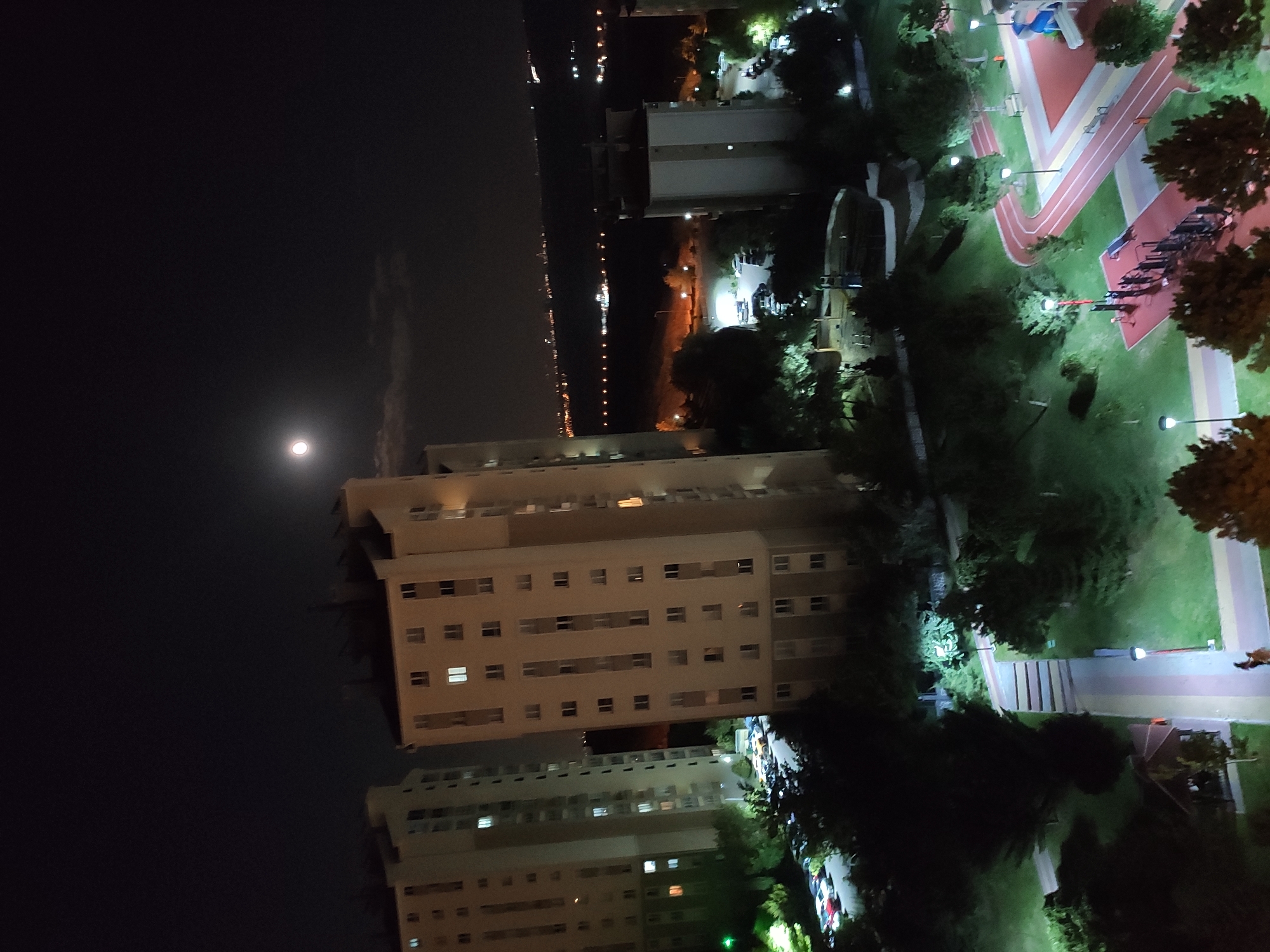 The image depicts a nighttime aerial view of a residential area in Etimesgut, Turkey. Two multi-story apartment buildings are prominent in the foreground, with numerous windows illuminated by interior lights. The sky above is dark, with a bright full moon positioned slightly left of center, partially obscured by a wispy cloud. In the distance, a line of city lights stretches across the horizon, suggesting a larger urban landscape. Below the buildings, well-lit park areas with walking paths, trees, and children's playground equipment are visible. Several cars are parked in designated lots near the apartment buildings. The overall scene conveys a sense of a peaceful, lit-up urban environment at night.
