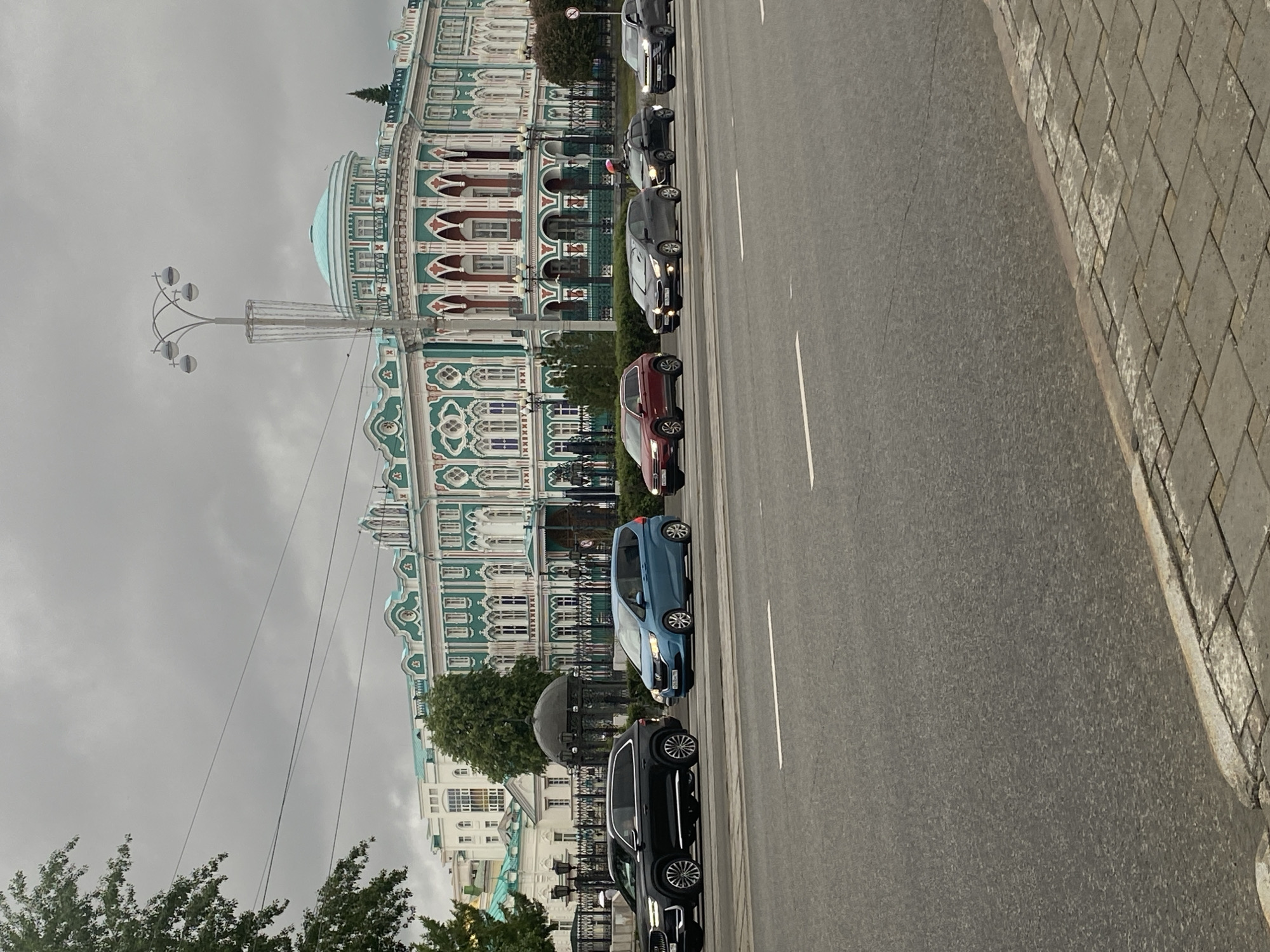 The image depicts a street scene in Yekaterinburg, Russia, featuring the Sevastyanov House as a prominent architectural landmark. A line of parked cars, including a blue hatchback and a red sedan, sits adjacent to the building along a multi-lane road. The road is marked with white lane dividers, and a sidewalk paved with gray brick runs along the right side of the frame. A decorative street light stands to the left of the frame near the building. The sky is overcast, suggesting a cloudy day.