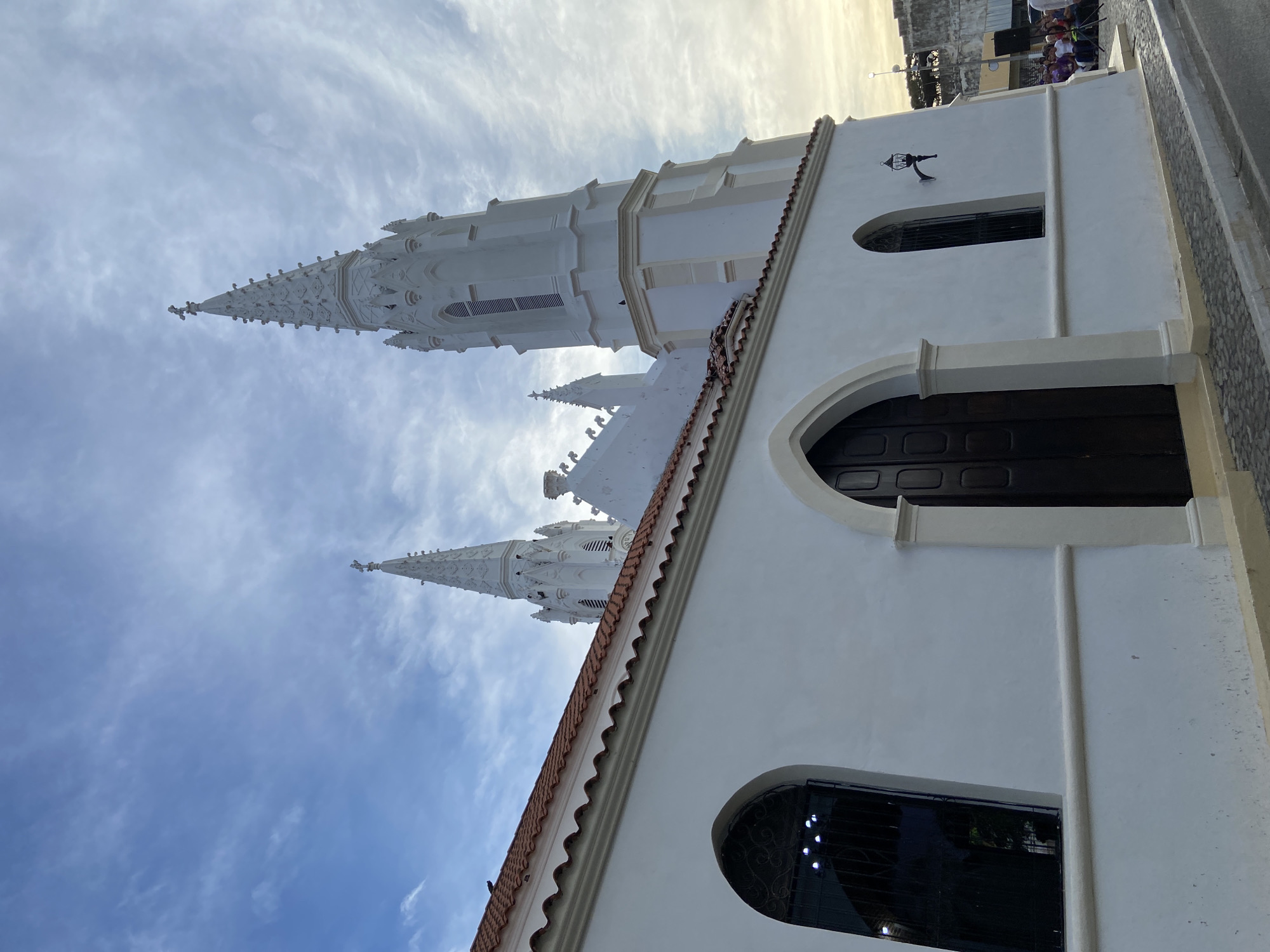 The image shows the Iglesia de San Juan Evangelista, a Roman Catholic church in Juan Griego, Venezuela. The angle is a low shot looking up, emphasizing the white church's architecture against a blue, partly cloudy sky. Visible are the church's spires, white walls, arched windows, and red-tiled roof. People are seen near the church.