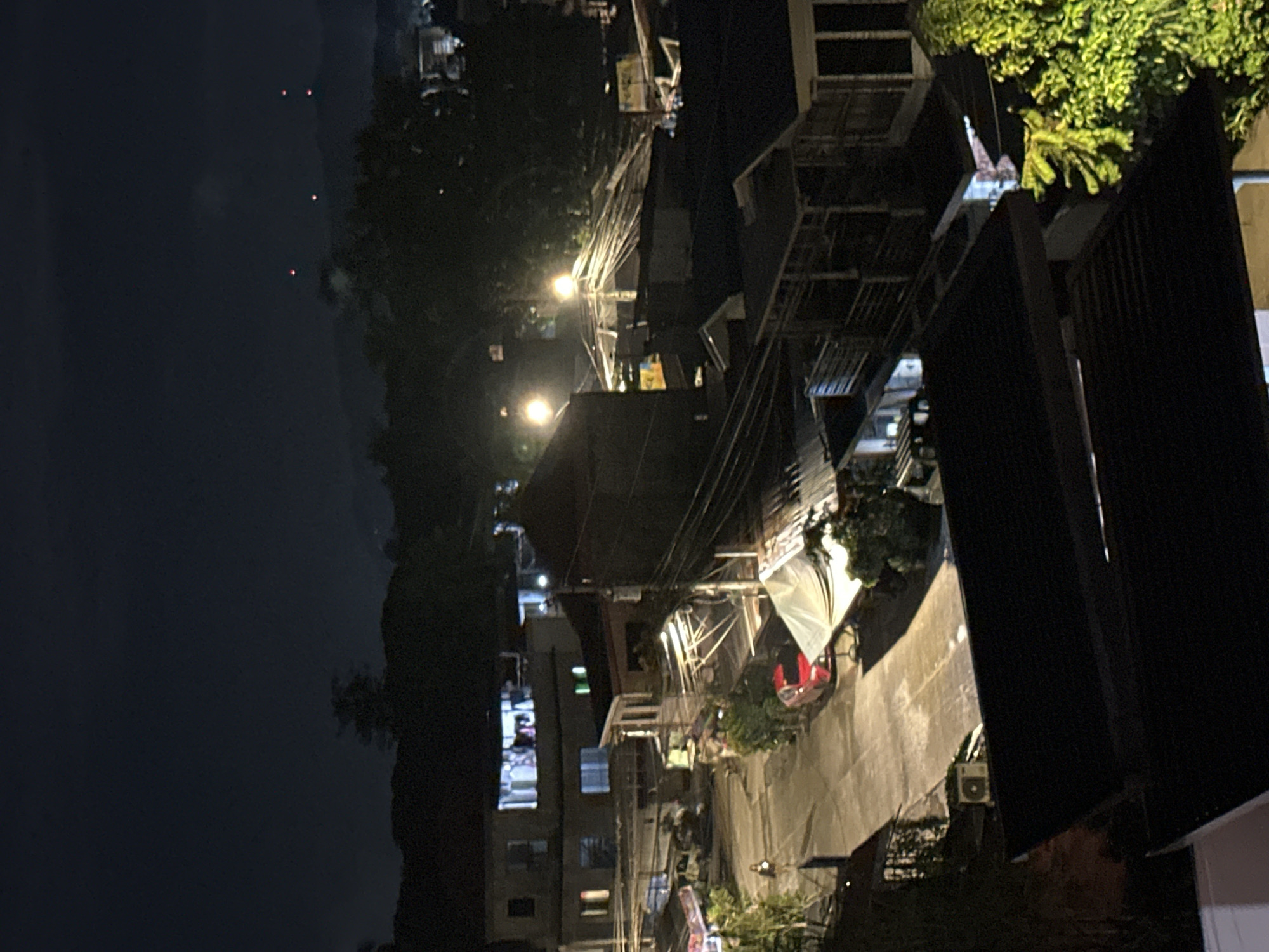 An elevated night-time view captures a street scene in a residential area of Davao City, Philippines. Buildings with multiple stories and various roof types, including corrugated metal and flat surfaces, are visible. Lights emanate from windows and street lamps, illuminating the paved street and sidewalks. A red car is parked on the street, and a person can be seen walking. Tents or tarps are set up over some structures, suggesting ongoing construction or temporary shelters. Electrical wires are strung between poles, a common sight in urban Philippines. In the background, distant mountains are silhouetted against the dark sky, with a few faint red lights visible, possibly indicating aircraft or distant signals. The overall impression is one of a typical urban residential neighborhood at night.