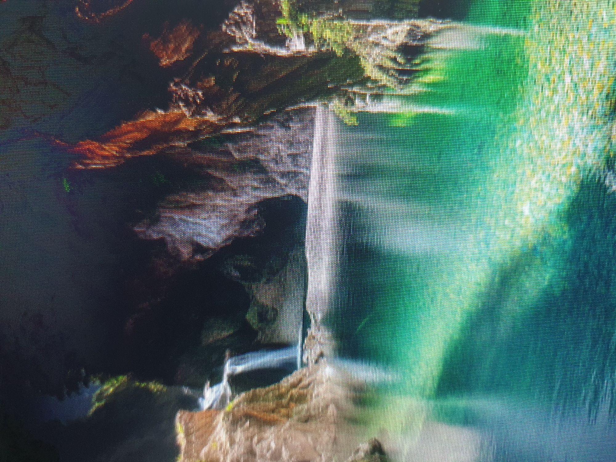 The image depicts a serene natural landscape featuring a body of clear, turquoise water surrounded by lush vegetation and rocky formations. A small waterfall cascades into the water on the left side of the frame. The water is shallow enough to reveal the rocky riverbed beneath, dotted with pebbles and small stones. Sunlight filters through the foliage, casting dappled shadows on the water's surface and the surrounding rocks. The scene evokes a sense of tranquility and natural beauty. There are no people or man-made structures visible in the image. The location is not identifiable as being in the city of Tashkent, Uzbekistan.
