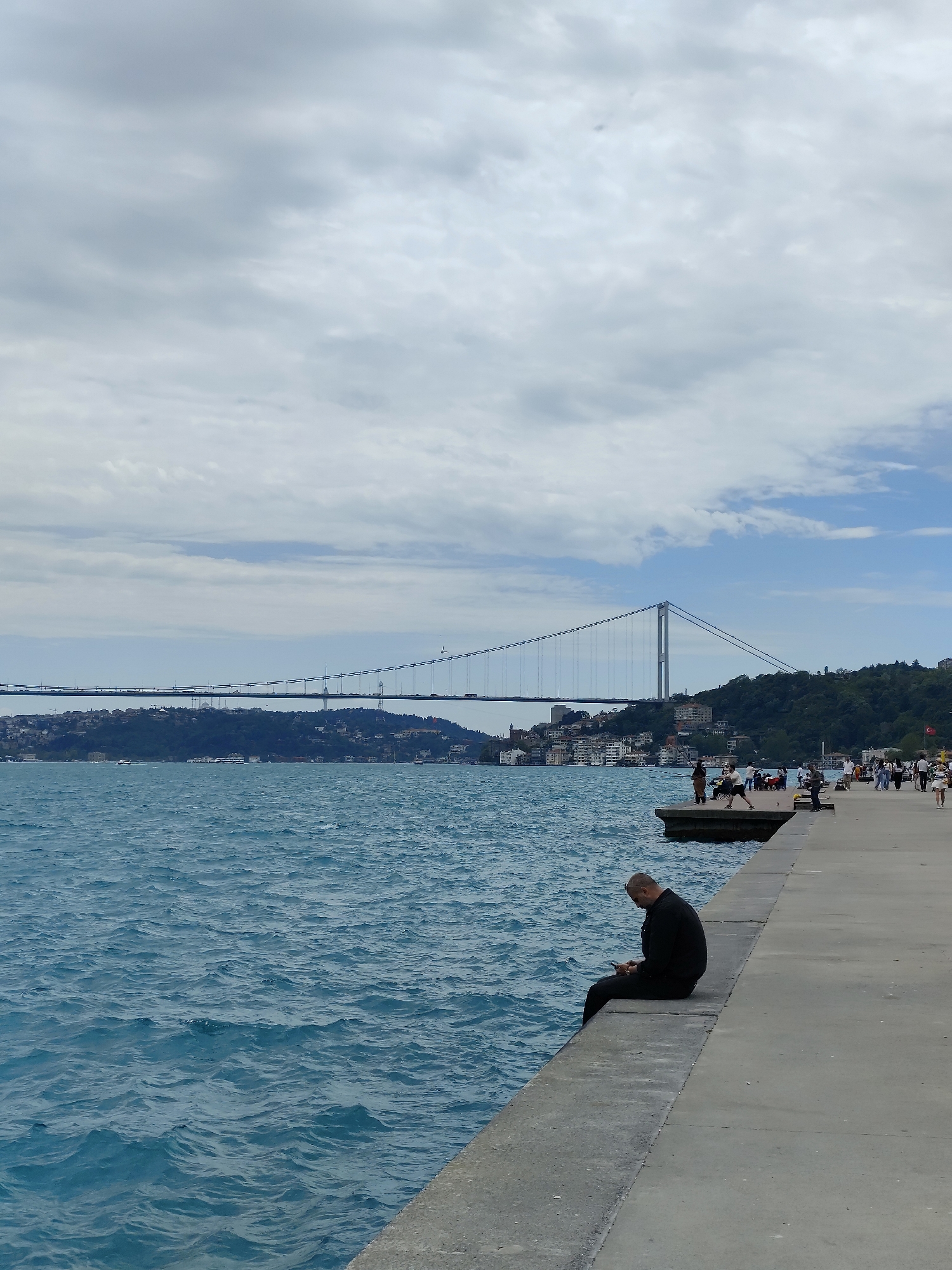 A man in dark clothing sits on the edge of a concrete quay, facing the Bosphorus strait in Sarıyer, Turkey. He appears to be using a mobile phone. The water is choppy, with waves visible. In the background, the Bosphorus Bridge spans the strait, connecting Europe and Asia. The shorelines on both sides of the strait are populated with buildings and green foliage. Several people are standing on the quay to the right. The sky is overcast with a mix of clouds and patches of blue.