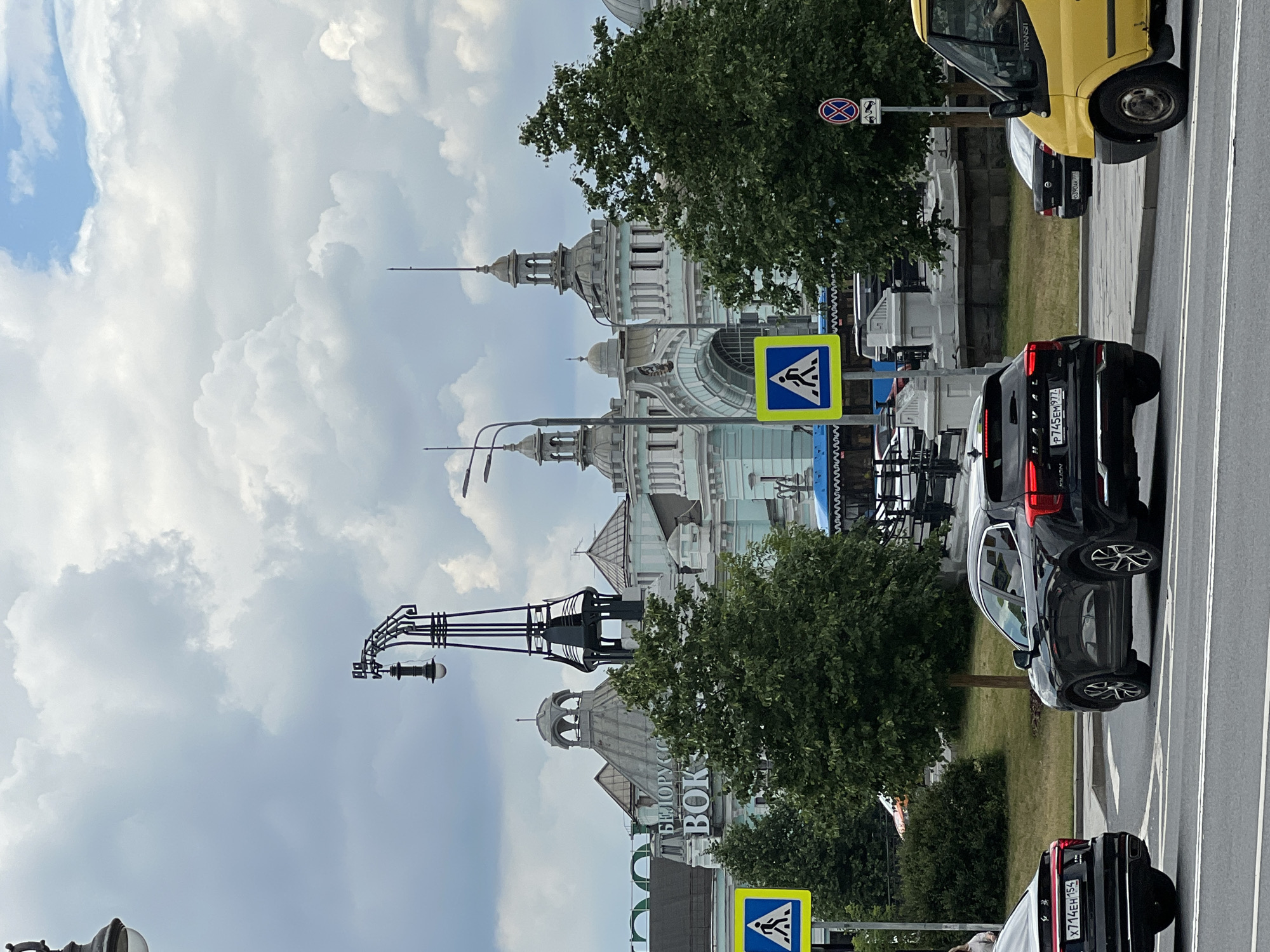 The image captures a view of the Belorussky railway station in Moscow, Russia, under a cloudy sky. In the foreground, a black SUV is stopped at a pedestrian crossing, with its brake lights illuminated. To the left of the SUV, another black car is also visible, partially obscured by the SUV. A yellow van is positioned to the right, with its front end in the frame.
The background features the distinctive architecture of the railway station, characterized by its domes, towers, and ornate facade. Trees line the area in front of the station, partially obscuring the lower levels. Two pedestrian crossing signs, one in the left portion of the image and another slightly to the right, are clearly visible, indicating designated areas for pedestrians to cross the road. Streetlights and traffic signs are also present. The overall scene depicts a typical urban environment with active traffic flow.