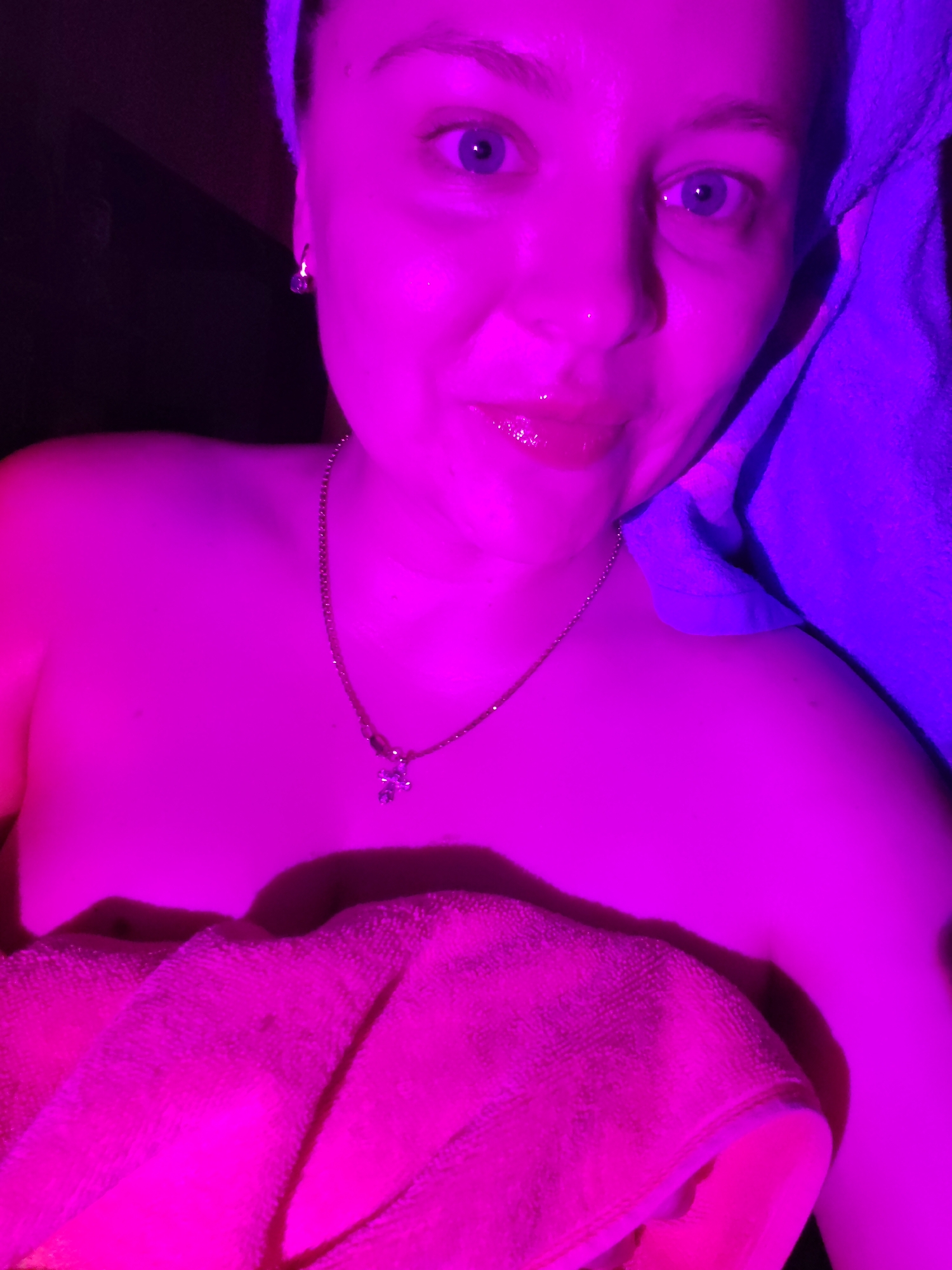 The image is a close-up, selfie-style photograph of a young woman with blue eyes and light skin. She is wearing a small silver necklace with a cross pendant. She has a pink towel wrapped around her upper body and a blue towel draped over her head and shoulder. The scene is illuminated by pink and purple lighting, creating a vibrant and somewhat surreal atmosphere. The background is dark and indistinct. No specific location in Izhevsk, Russia, can be identified from the image.