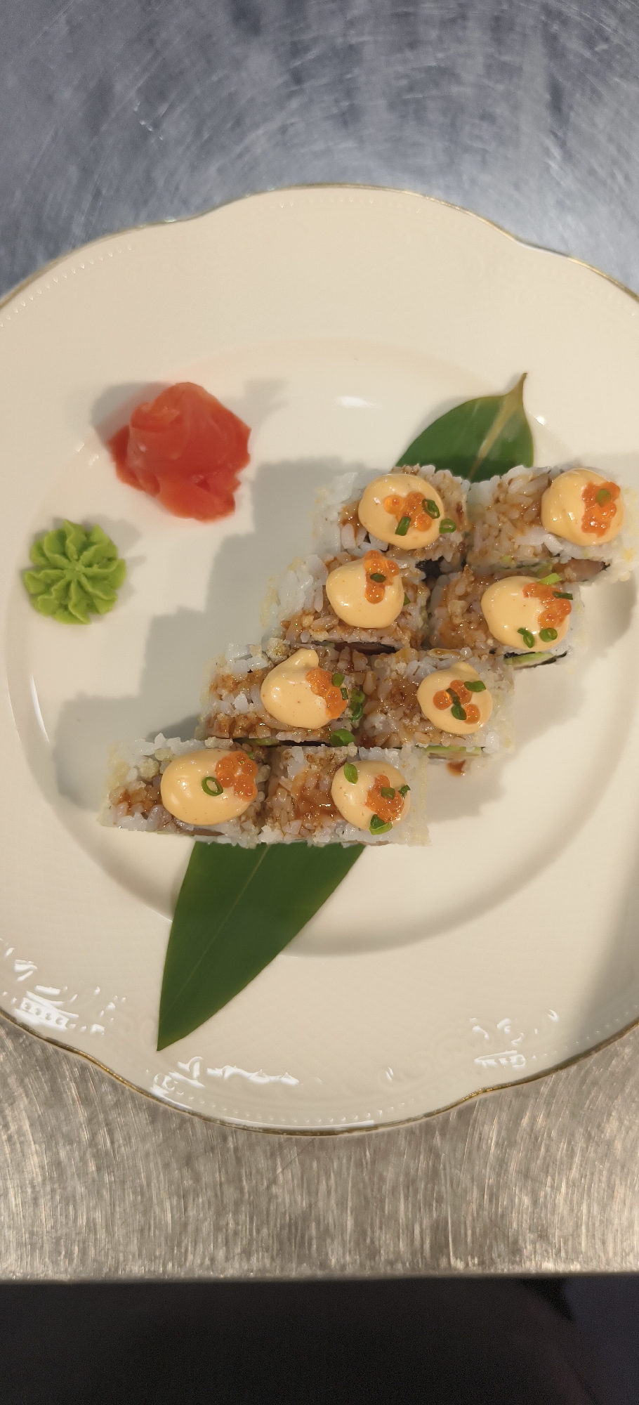 The image shows a plate of sushi, accompanied by a leaf, wasabi, and pickled ginger, resting on a stainless steel countertop. The sushi consists of a roll cut into several pieces, topped with a creamy sauce and sprinkled with orange roe and chopped green onions. The plate is white with a textured border and gold trim. The wasabi is shaped like a small flower. The countertop has a brushed, metallic texture. The image was taken in Yerevan, Armenia.
