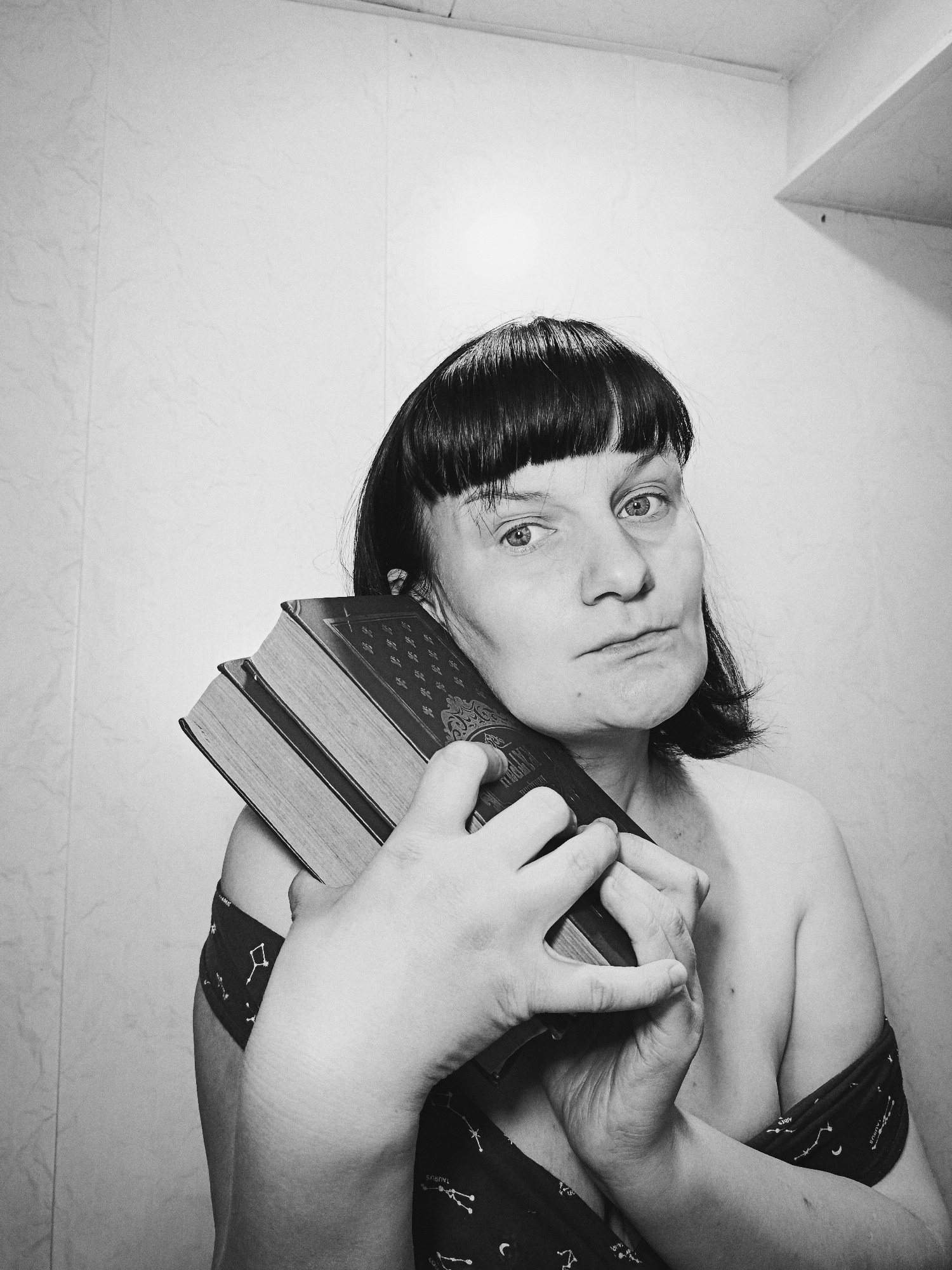 A black and white medium shot photograph features a person holding three thick books. The person, with a serious expression, has short, dark hair and is partially bare-shouldered, wearing a dark garment patterned with astrological constellations. The background is a plain, light-colored wall, possibly tiled. A small shelf is visible in the upper right corner. The location is not determinable from the image alone.