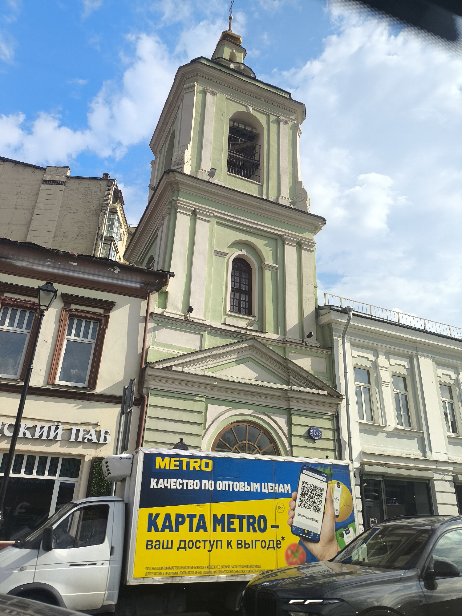 The image is a street scene, likely taken in Moscow, Russia, depicting a pale green and white multi-story bell tower of a Russian Orthodox church, with a gold dome and cross at its peak. The church is adjacent to other buildings of varying heights and architectural styles, including a building with a sign in Cyrillic script ("ский паб"). A yellow and blue Metro Cash & Carry truck is partially obscuring the base of the church, featuring an advertisement in Russian for a Metro card. A gray BMW automobile is parked in front of the truck. The sky is blue with scattered white clouds. A street sign indicating "50/2" is seen on one of the buildings.