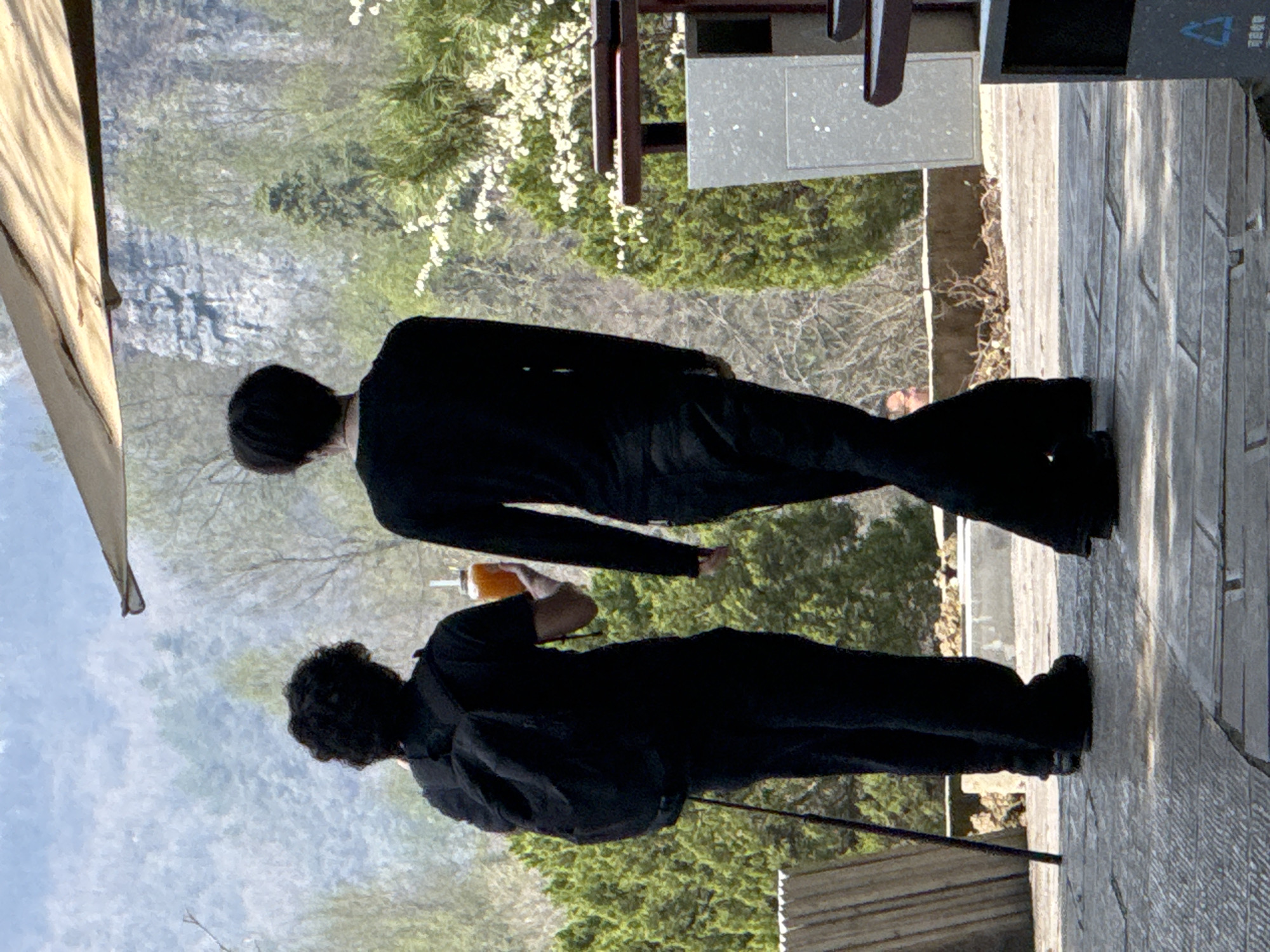 The image depicts two individuals walking away from the viewer on a paved pathway in an outdoor setting within Wulingyuan, China. The person on the left, with dark, curly hair, wears a dark short-sleeved top, dark pants, and a dark backpack. This individual holds a clear glass jar containing an orange liquid and a white straw in their right hand, and a dark, slender stick or monopod extends from their left hand to the ground. The second individual, positioned to the right and slightly ahead, wears a dark long-sleeved top and dark, possibly flared, pants, and has dark, straight hair.
The paved ground consists of rectangular, light-colored tiles. The immediate background features dense green foliage, including shrubs and trees, with patches of white flowering bushes on the right. In the mid-ground and distance, rugged grey rock formations and hazy, tree-covered mountains are visible, consistent with the Wulingyuan landscape. A light-colored canvas umbrella canopy is partially visible at the top center-right. On the far left, a vertical wooden slatted structure is present. On the far right, a grey rectangular utility-like object is visible, and a portion of a grey waste receptacle with a blue recycling symbol is at the bottom right. A faint, blurred reflection or a partially obscured person's head is visible behind the right individual's leg.