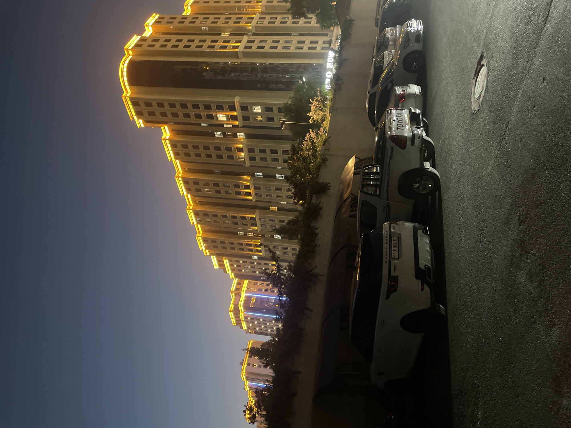 The image depicts a nighttime scene in Dushanbe, Tajikistan, featuring several modern apartment buildings illuminated with yellow and blue lights against a twilight sky. In the foreground, a parking lot is visible with several cars, including a white Range Rover with the license plate "1010EU10", a white Toyota pickup truck, and other sedans. The buildings are tall and multi-story, with a contemporary architectural style, and the lights accentuating their edges. Trees and shrubs line the area between the buildings and the parking lot. A sign on the building facade reads "ETRO HOM". The overall impression is one of urban development and modern living in Dushanbe.