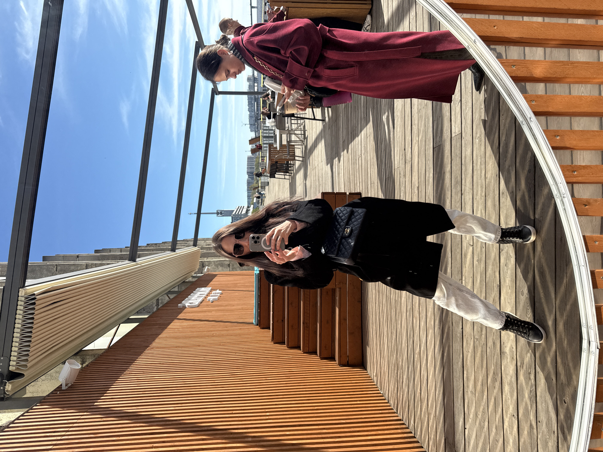 The image shows an outdoor scene in what appears to be a rooftop or terrace area in Saint Petersburg, Russia, as indicated by the city buildings in the background. A woman is standing on a wooden deck, taking a photo with her smartphone. She is wearing a black coat, light-colored pants, and black shoes, carrying a black quilted handbag. Behind her, there are wooden steps and a metal structure, possibly part of the building's architecture or a sunshade. Another person is visible wearing a red coat and seems to be looking down. There are tables and chairs in the background, suggesting a café or restaurant setting. The day appears to be sunny with a blue sky. A decorative oval frame is partly visible on the right side of the image.